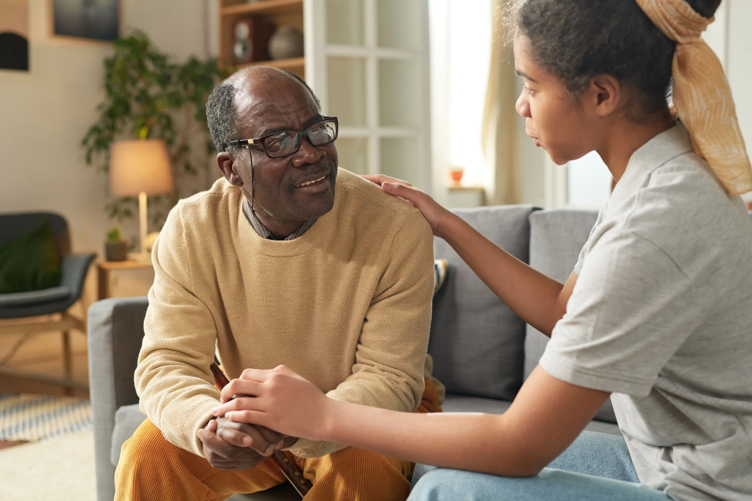 An elderly man talking to a young woman in a living room, with the woman placing her hand on the man's shoulder and holding his hands.
