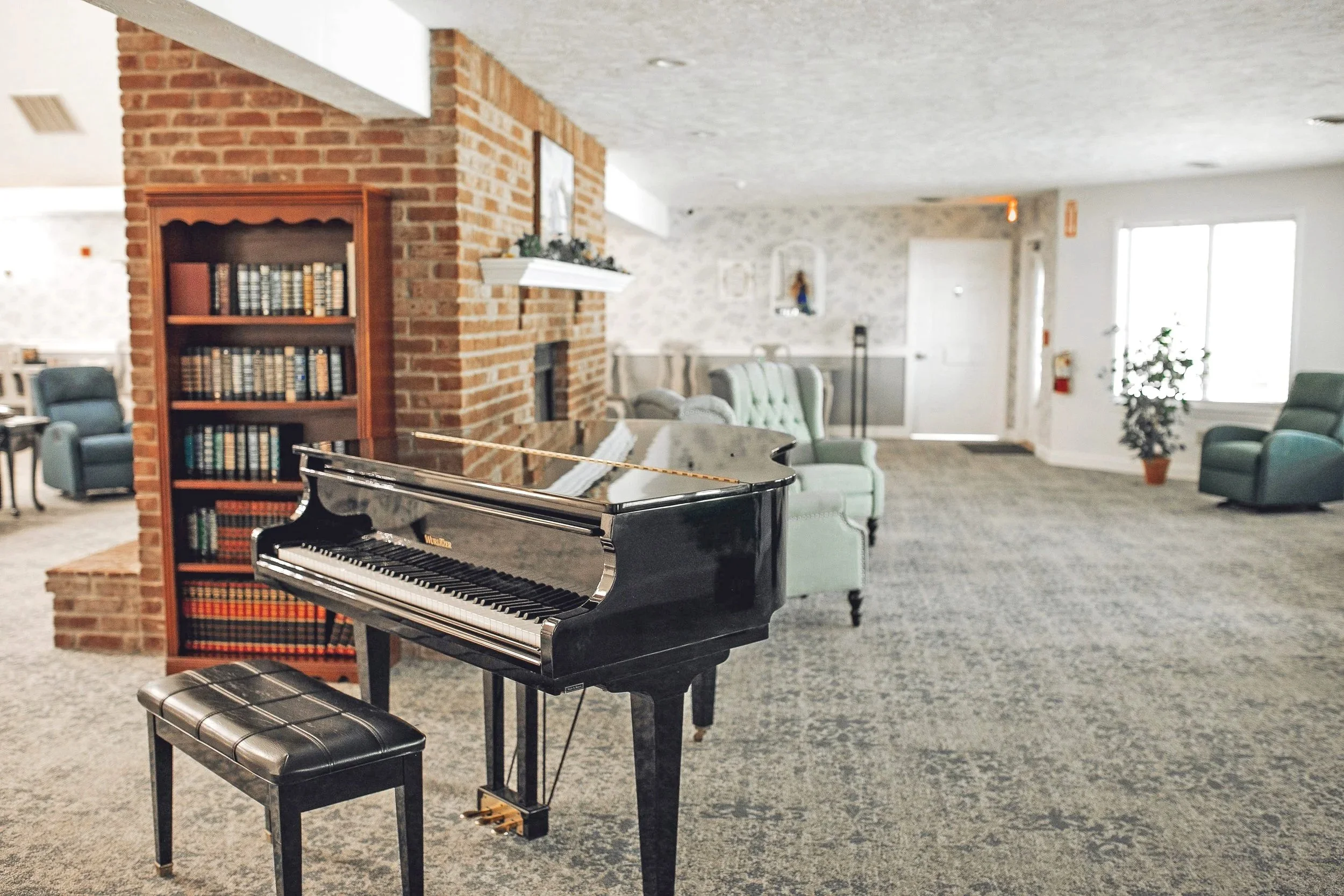 A black grand piano with a matching piano bench in a cozy, well-lit lounge or waiting area with green armchairs, a brick fireplace, a bookshelf, and potted plants.