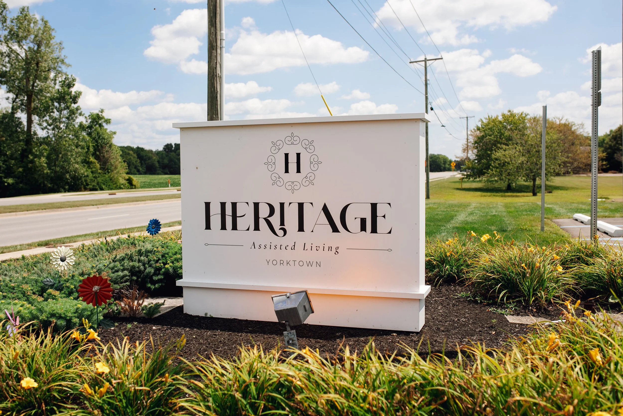 Sign for Heritage Assisted Living in Yorktown, placed in a landscaped area with flowers and bushes, near a road and utility poles under a partly cloudy sky.