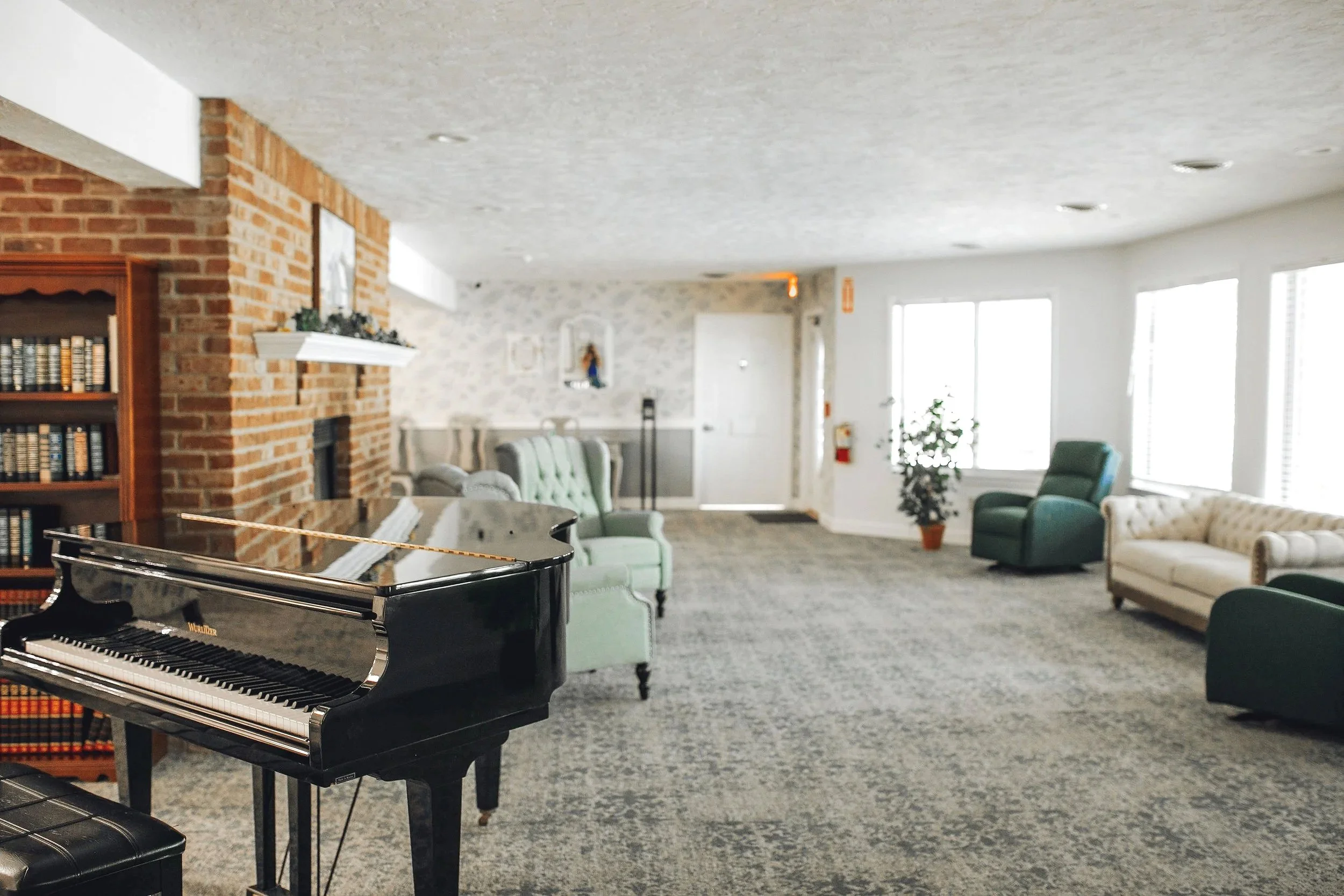 Interior of a spacious lounge or music room with a black grand piano, green and beige armchairs, a grey patterned carpet, a brick fireplace, bookshelf, and large windows letting in natural light.