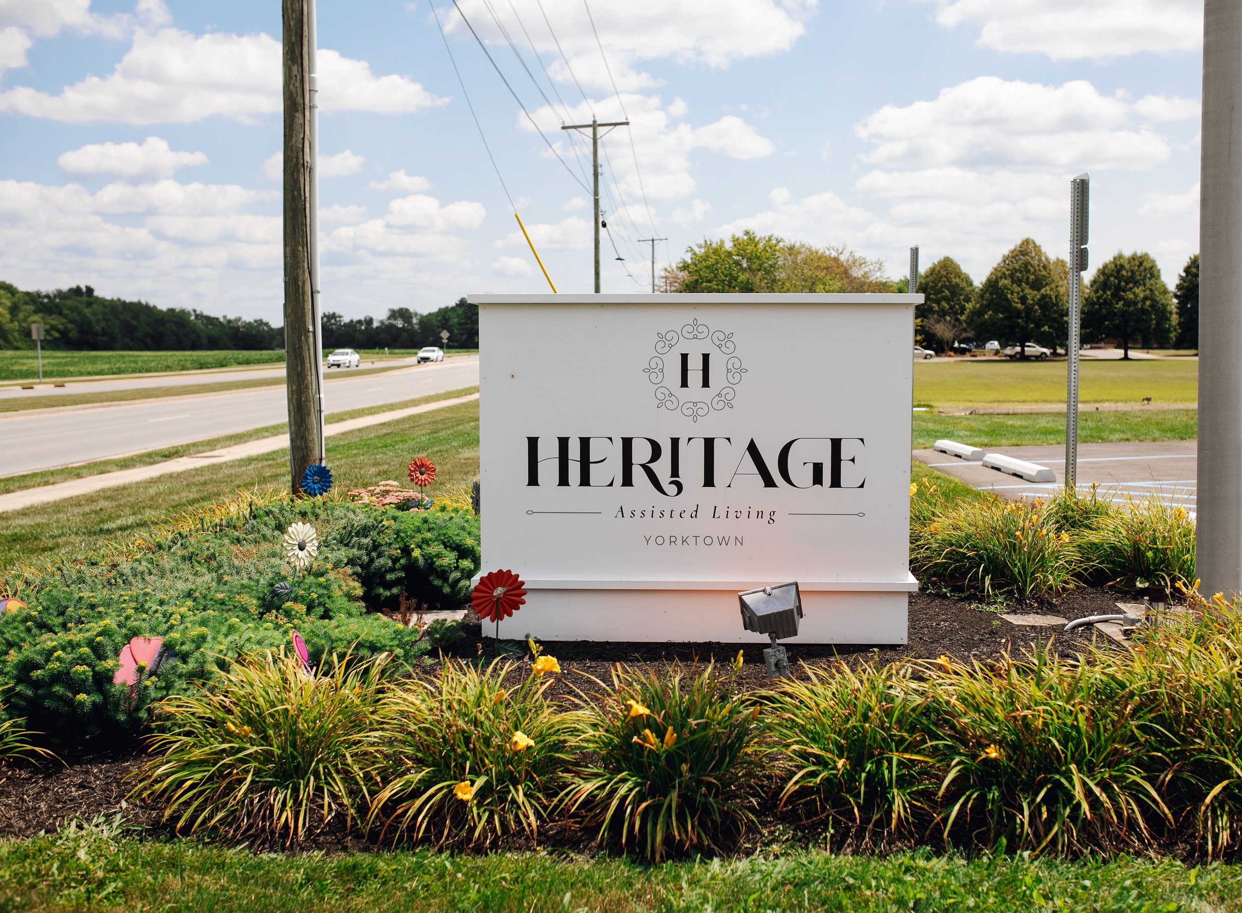 Sign for Heritage Assisted Living in Yorktown, surrounded by colorful flowers on a grassy area, with a road and parking lot in the background under a partly cloudy sky.