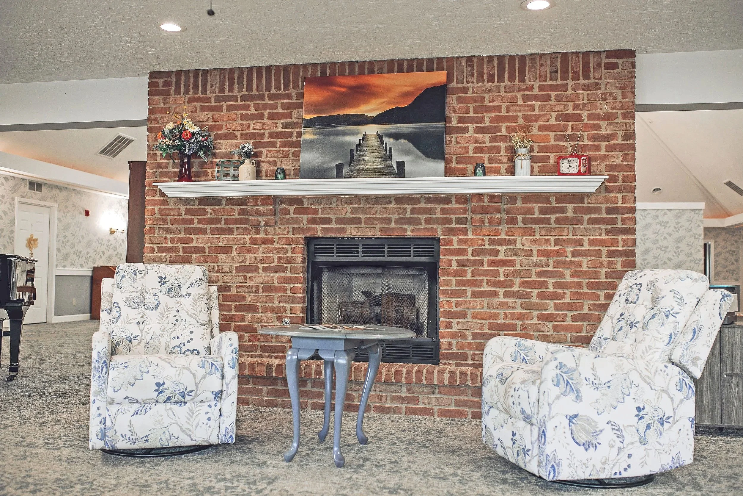 Living room with a brick fireplace, two floral armchairs, a small blue table, and a painting of a dock at sunset above the mantel. Decor includes flowers, vases, and a clock on the shelf.