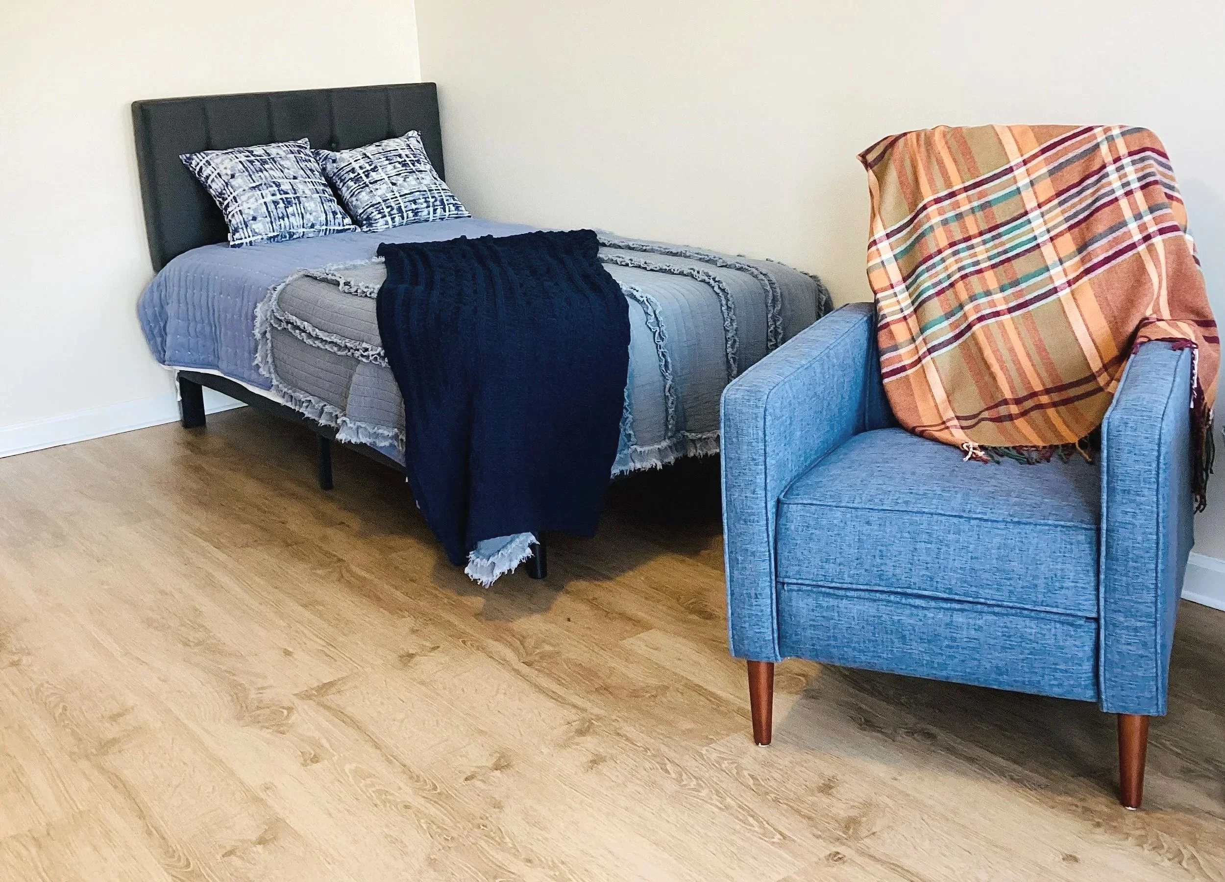 A bedroom with a bed against a white wall, dressed with gray bedding and two matching pillows, a dark blue throw blanket, and another blanket at the foot. Next to the bed is a blue armchair with a rolled arm, wooden legs, and an orange and yellow plaid blanket draped over it.