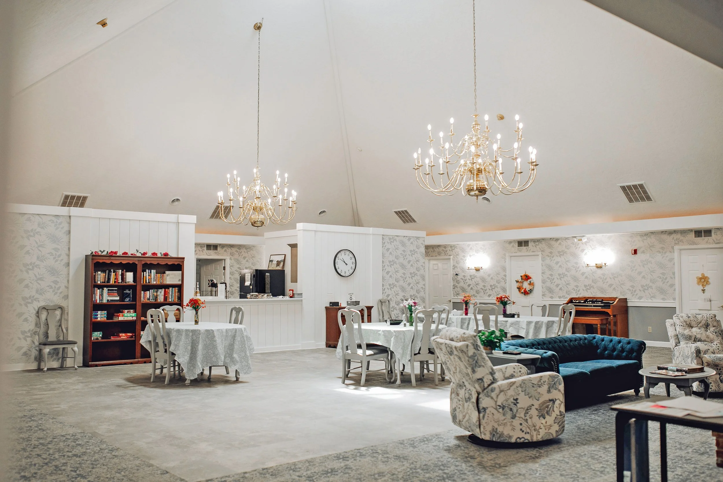 A spacious, well-lit community lounge with white walls, floral wallpaper, and elegant chandeliers. The room features white round tables with chairs, a floral wreath on the wall, a bookshelf, armchairs, a velvet sofa, a piano, and multiple flower vases.