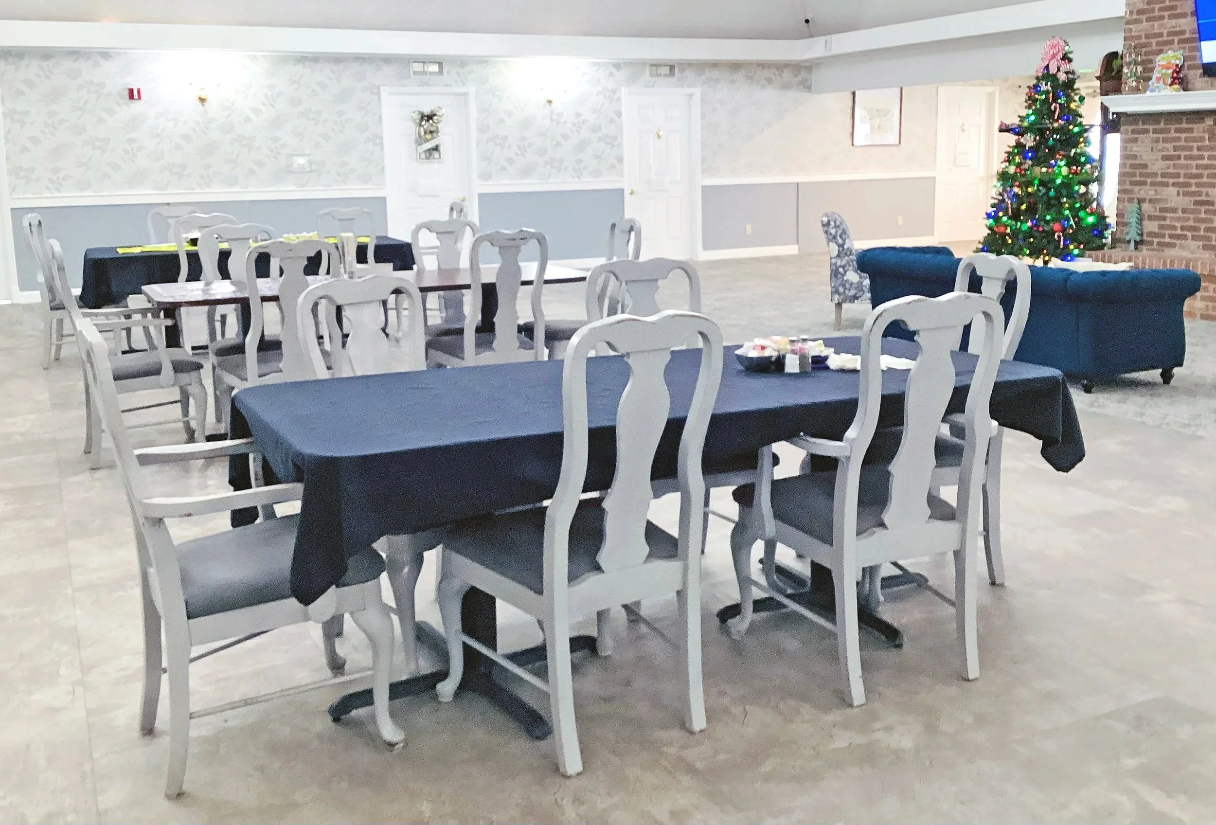 A large decorated Christmas tree with colorful lights and ornaments, in a spacious room with white walls, chairs with white frames and dark cushions, and tables covered with dark tablecloths, some with wrapped gifts on top.