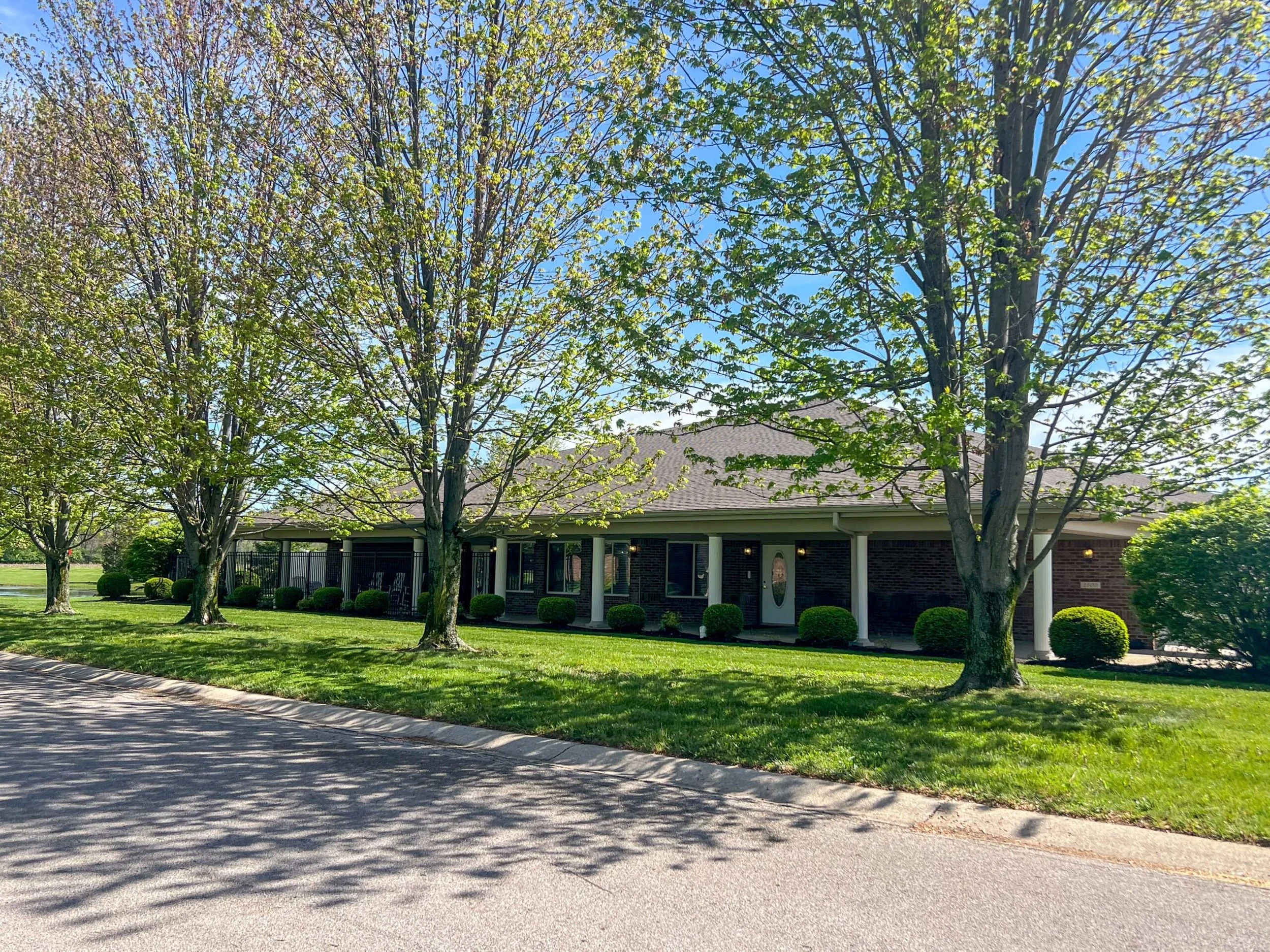 A single-story brick building with a front porch, surrounded by green lawn and neatly trimmed bushes, with large trees casting shadows on the sidewalk under a bright blue sky.