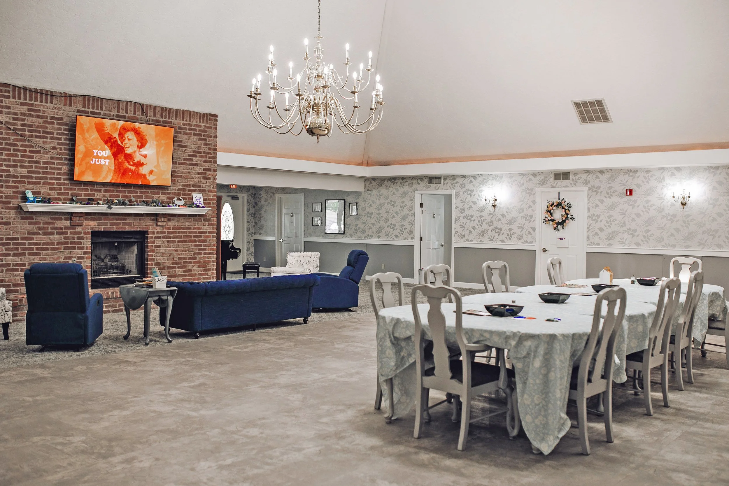 Event space with a brick fireplace, wall-mounted TV, blue and white seating, and a large dining table with white tablecloths, set with bowls and utensils, chandeliers, and decorative wall wallpaper.