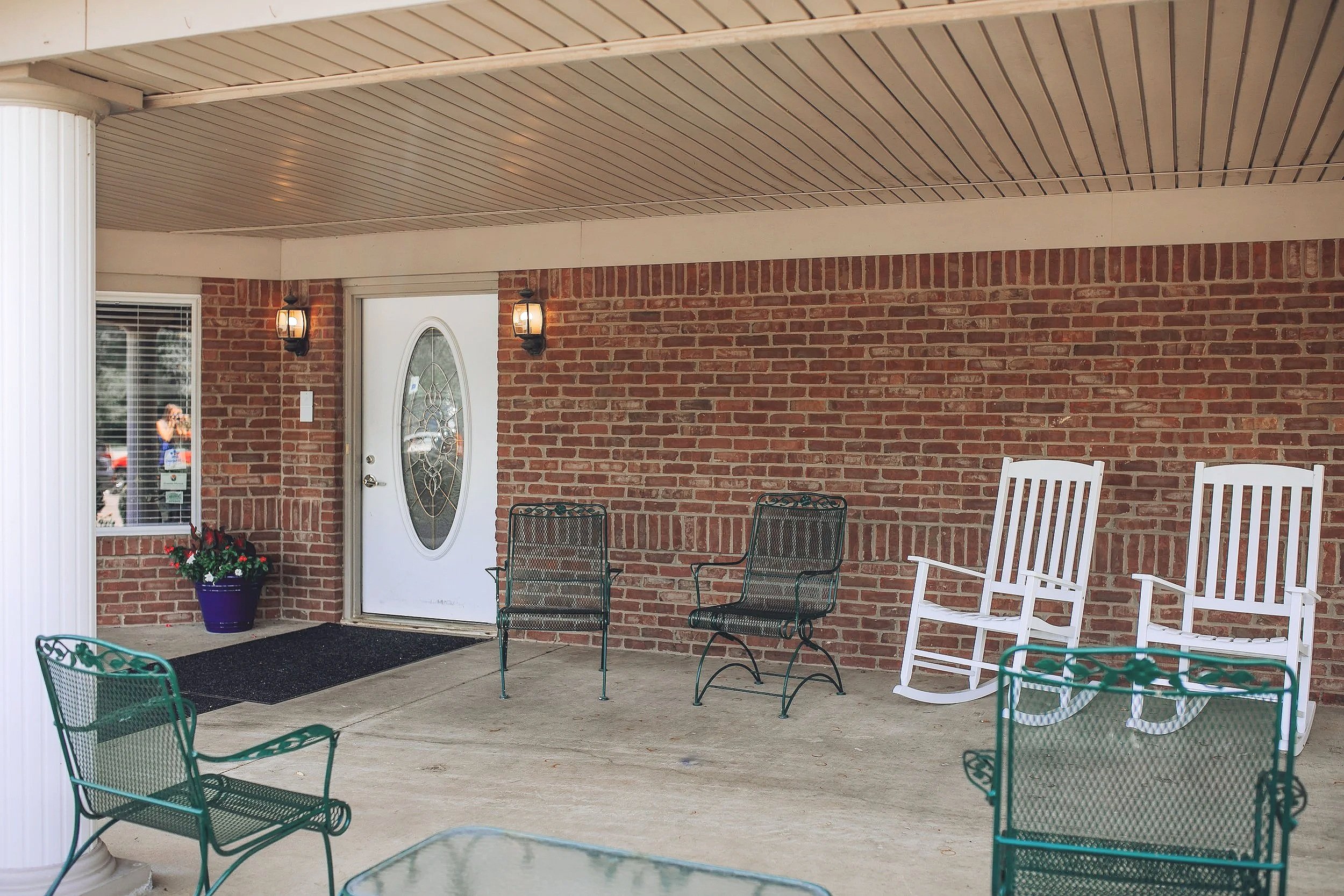 Porch with brick wall, white door with oval glass, and outdoor furniture including green metal chairs, white rocking chairs, a black doormat, and a flower pot with red flowers beneath a window.