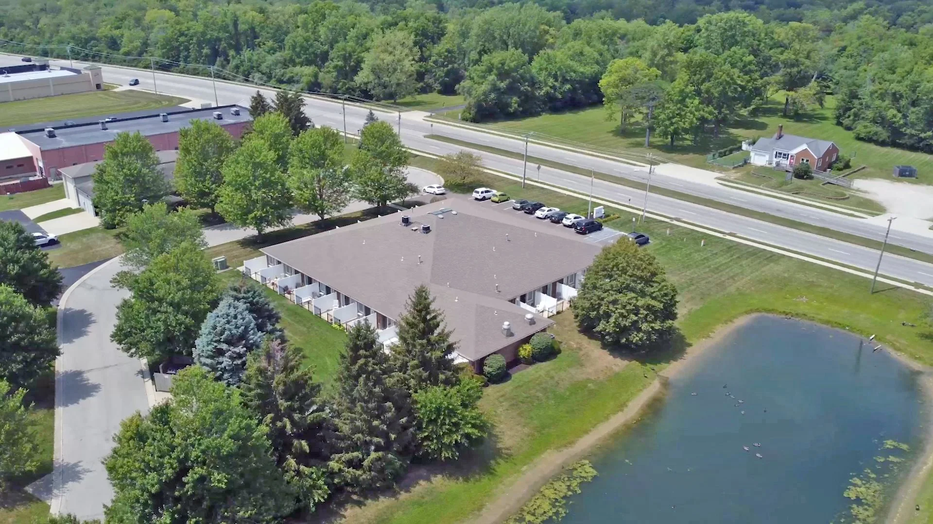 An aerial view of a residential area featuring a pond, a motel with multiple rooms, a parking lot with several cars, and a road with minimal traffic, surrounded by dense green trees and lawns.
