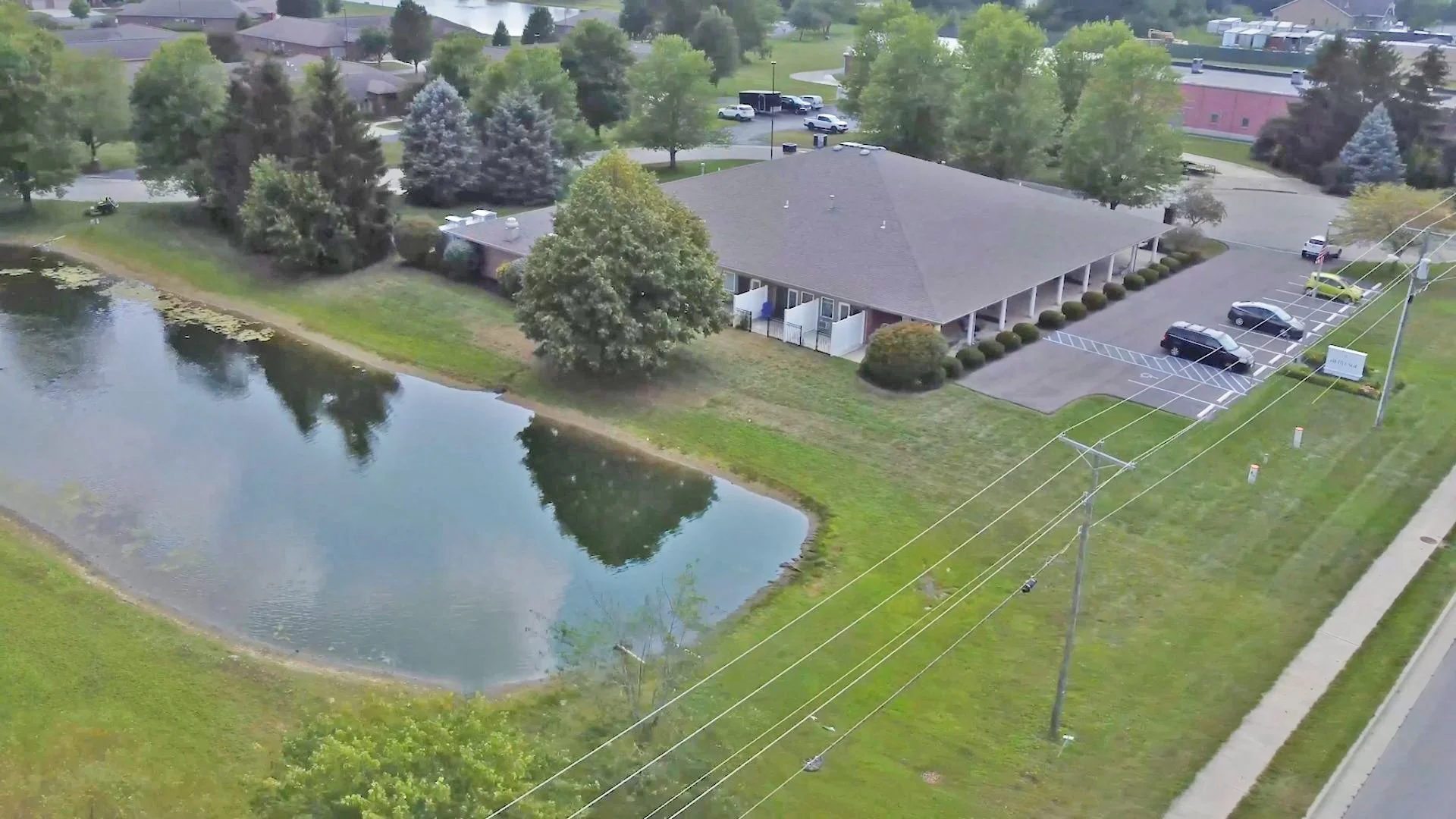 An aerial view of a building with a parking lot, trees, a pond, and power lines.
