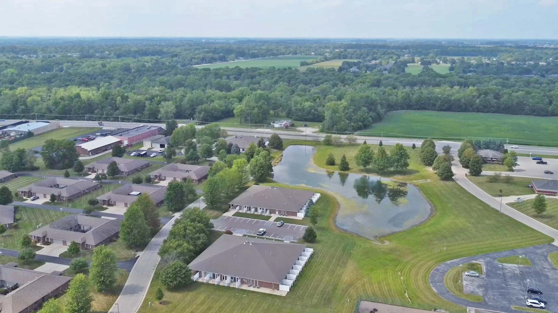 Aerial view of a suburban area with houses, a pond, parking lots, a highway, and lush green trees and fields in the background.