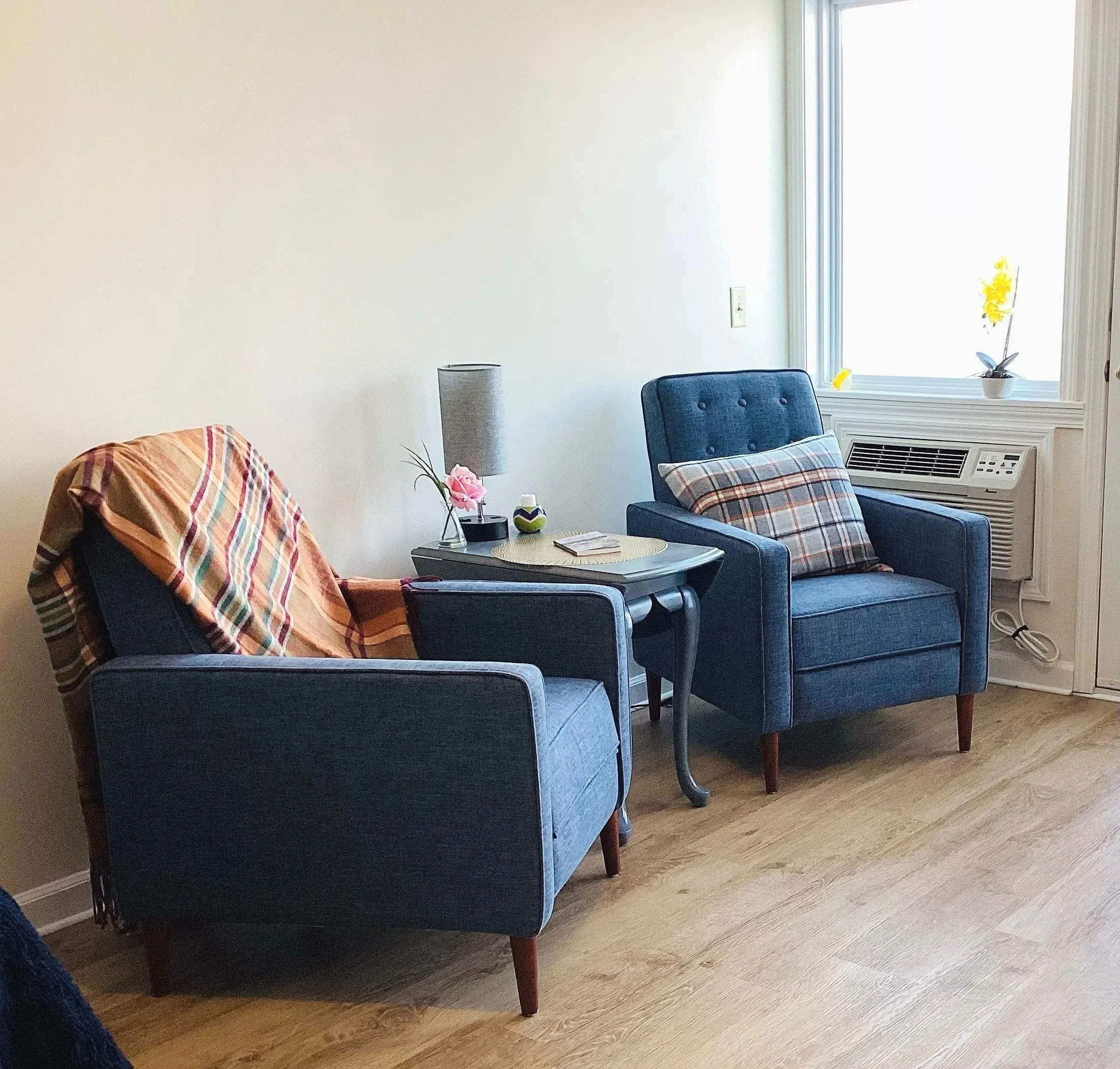 A cozy living room corner with two blue armchairs, a small side table with a lamp, flowers, and books, next to a window with yellow flowers on the windowsill.
