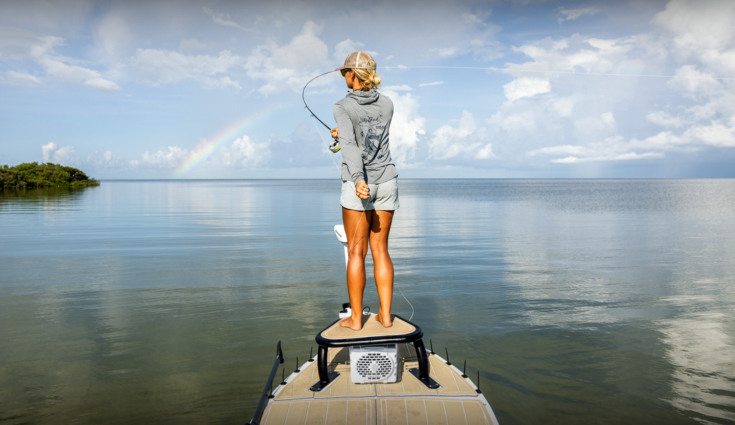 A woman standing on a boat fishing in a calm body of water with a rainbow in the background.