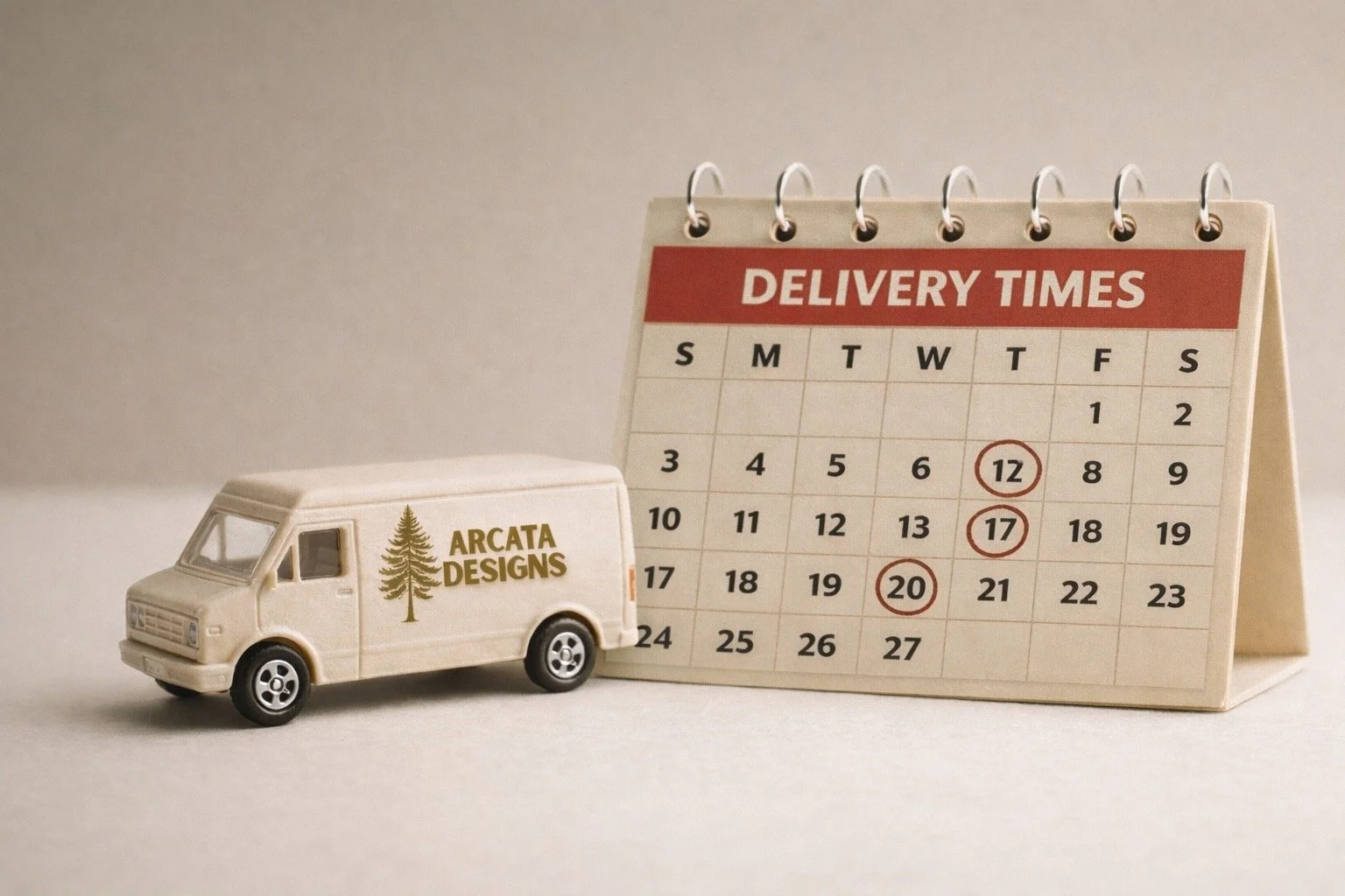 A miniature cream-colored delivery van with the Arcata Designs logo sits beside a large desk calendar labeled “Delivery Times,” with several dates circled in red, against a soft neutral background.