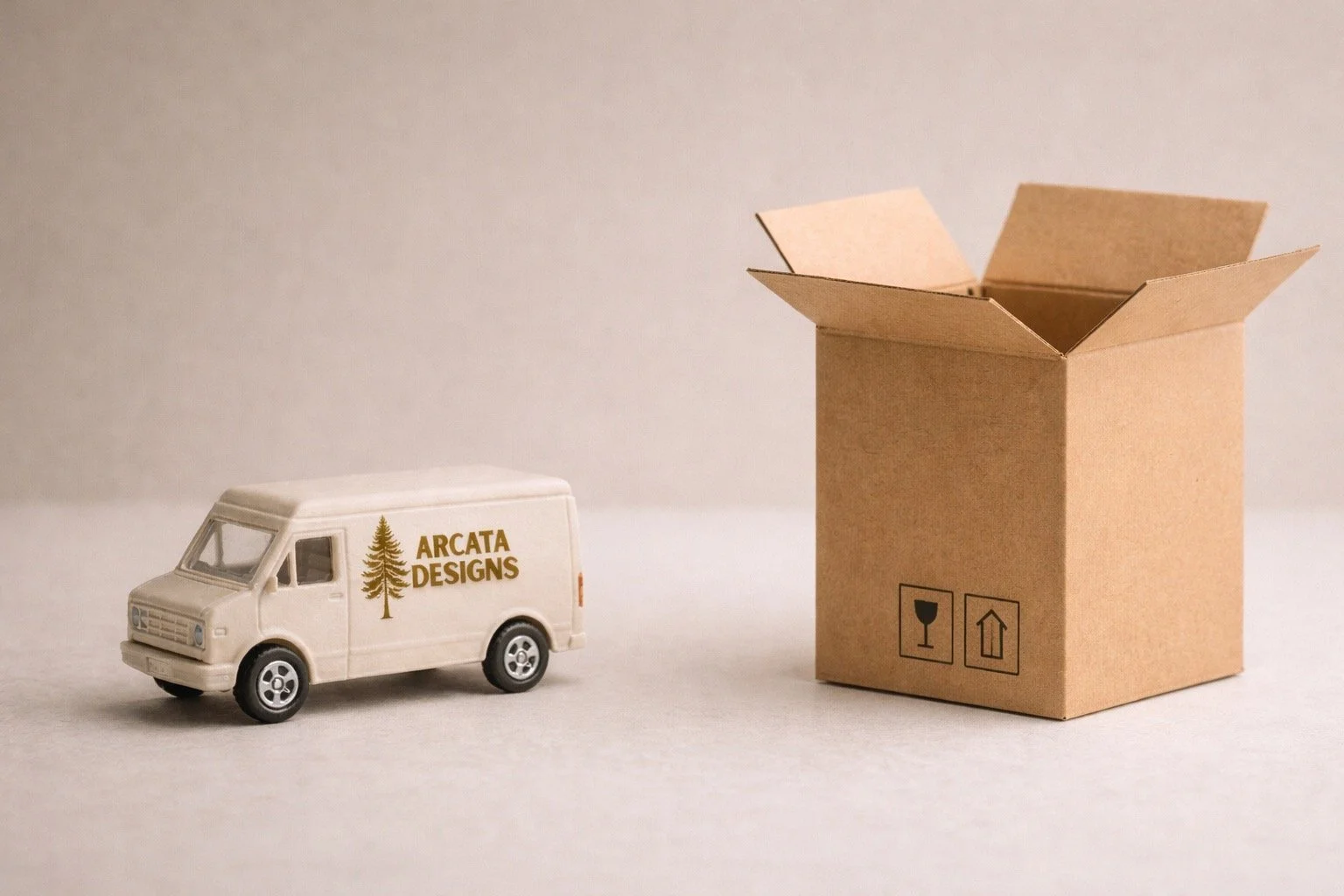 A miniature cream-colored delivery van with the Arcata Designs logo sits beside an open cardboard box with handling symbols, set against a soft neutral background.