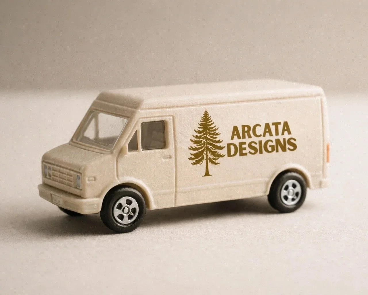 A miniature cream-colored delivery van with the Arcata Designs logo, a stylised evergreen tree and text, printed on its side, set against a soft neutral background.