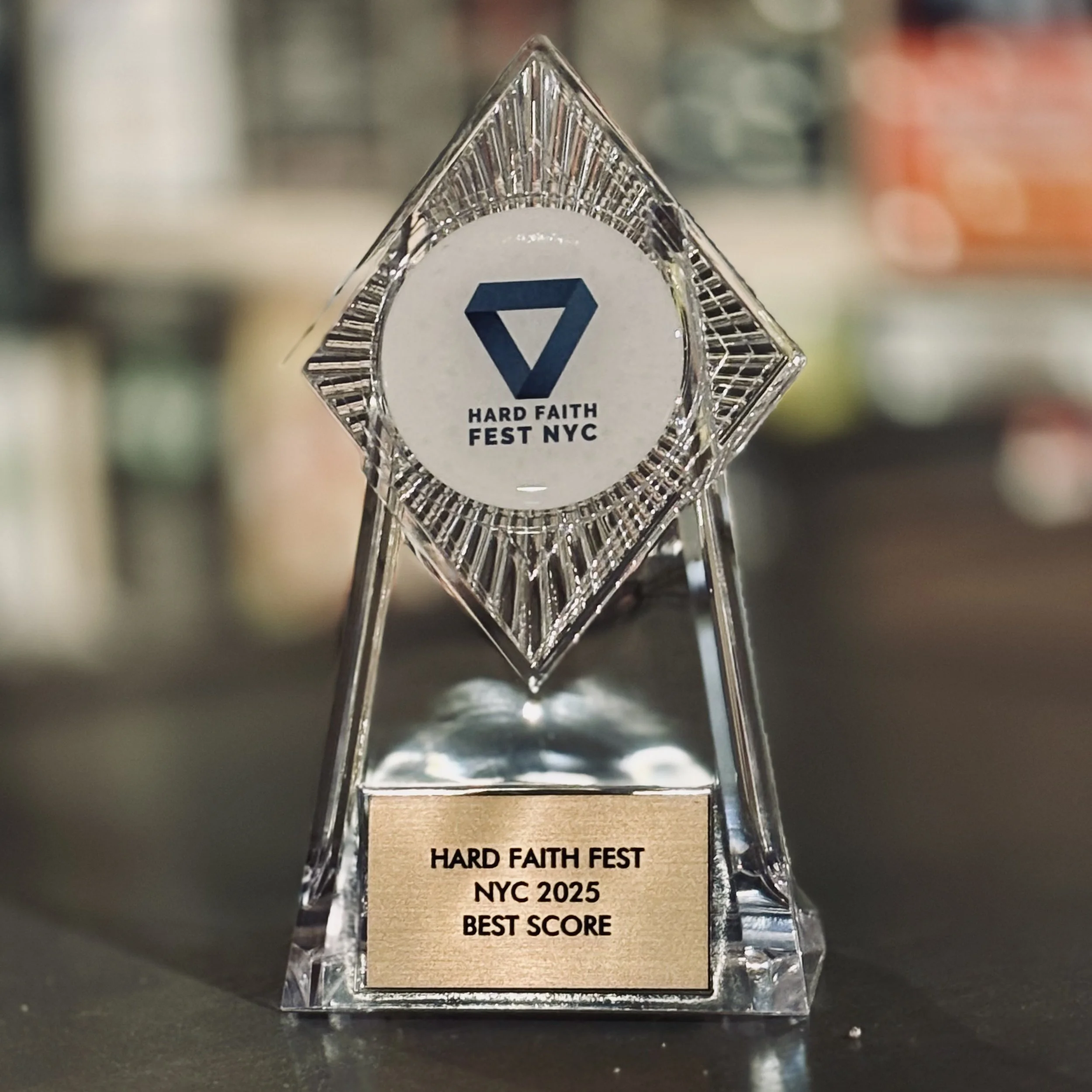 A glass award with a square base, featuring a gold plaque that reads 'HARD FAITH FEST NYC 2025 BEST SCORE' and a tilted diamond-shaped top with a circular emblem displaying a blue triangle and the text 'HARD FAITH FEST NYC'.