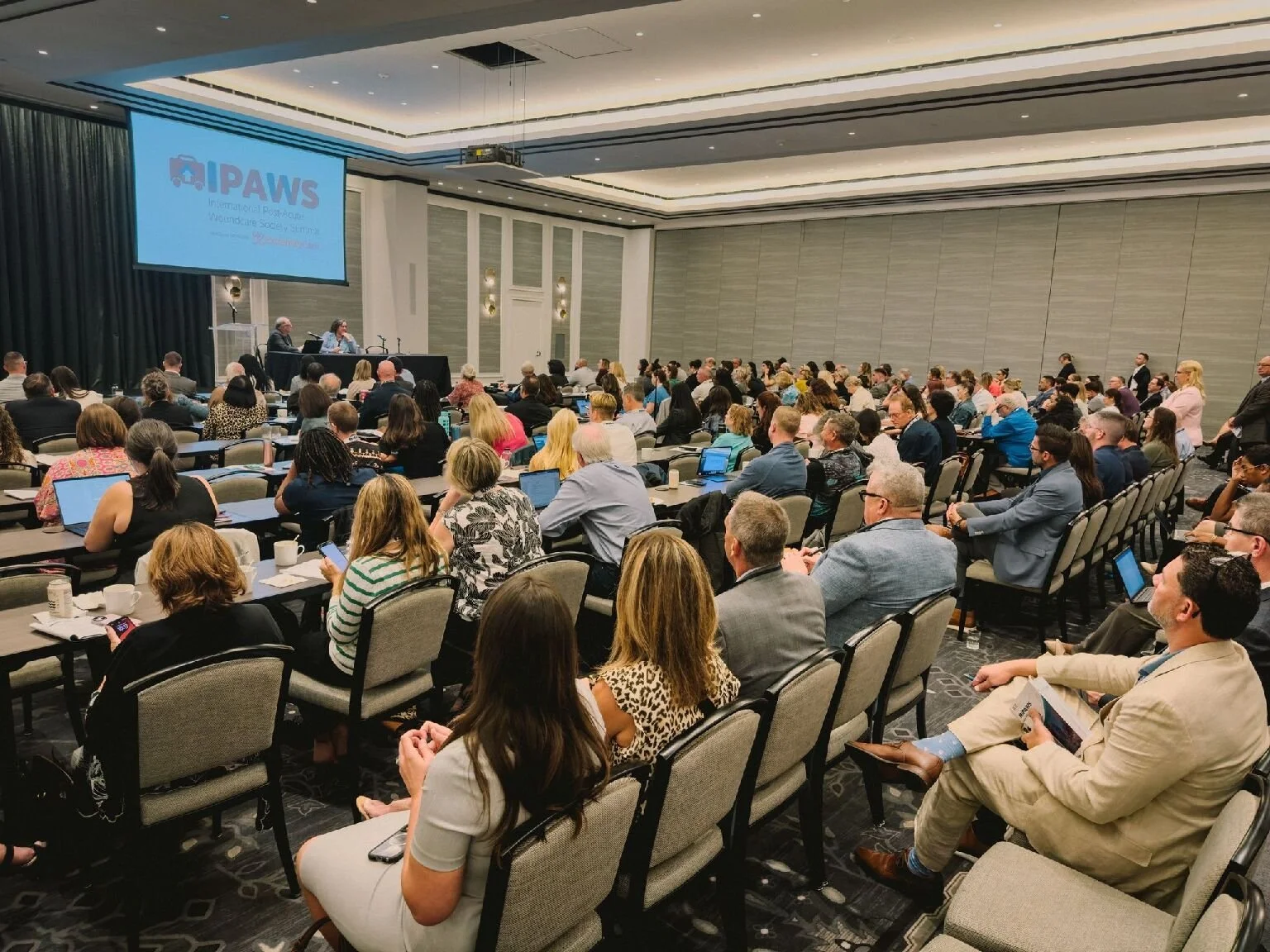 A conference room filled with attendees seated at tables, facing a stage with a large screen displaying the logo for PAWS.