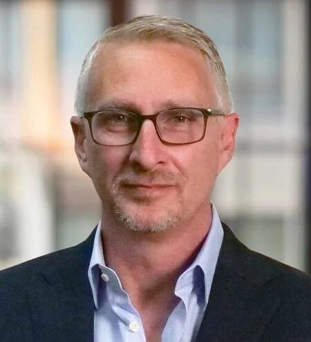 A middle-aged man with short gray hair, glasses, and a light beard, wearing a dark suit jacket and a light blue dress shirt, standing indoors with blurred windows in the background.