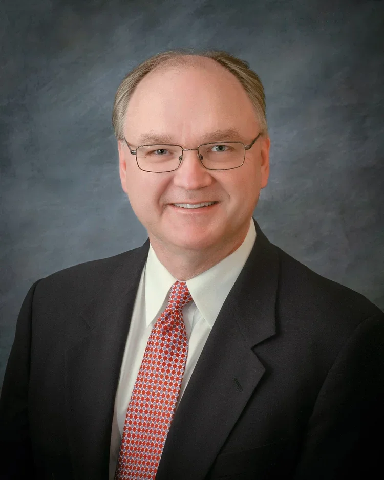 A professional headshot of a smiling middle-aged man with glasses, wearing a dark suit, white shirt, and red patterned tie, against a gray textured background.