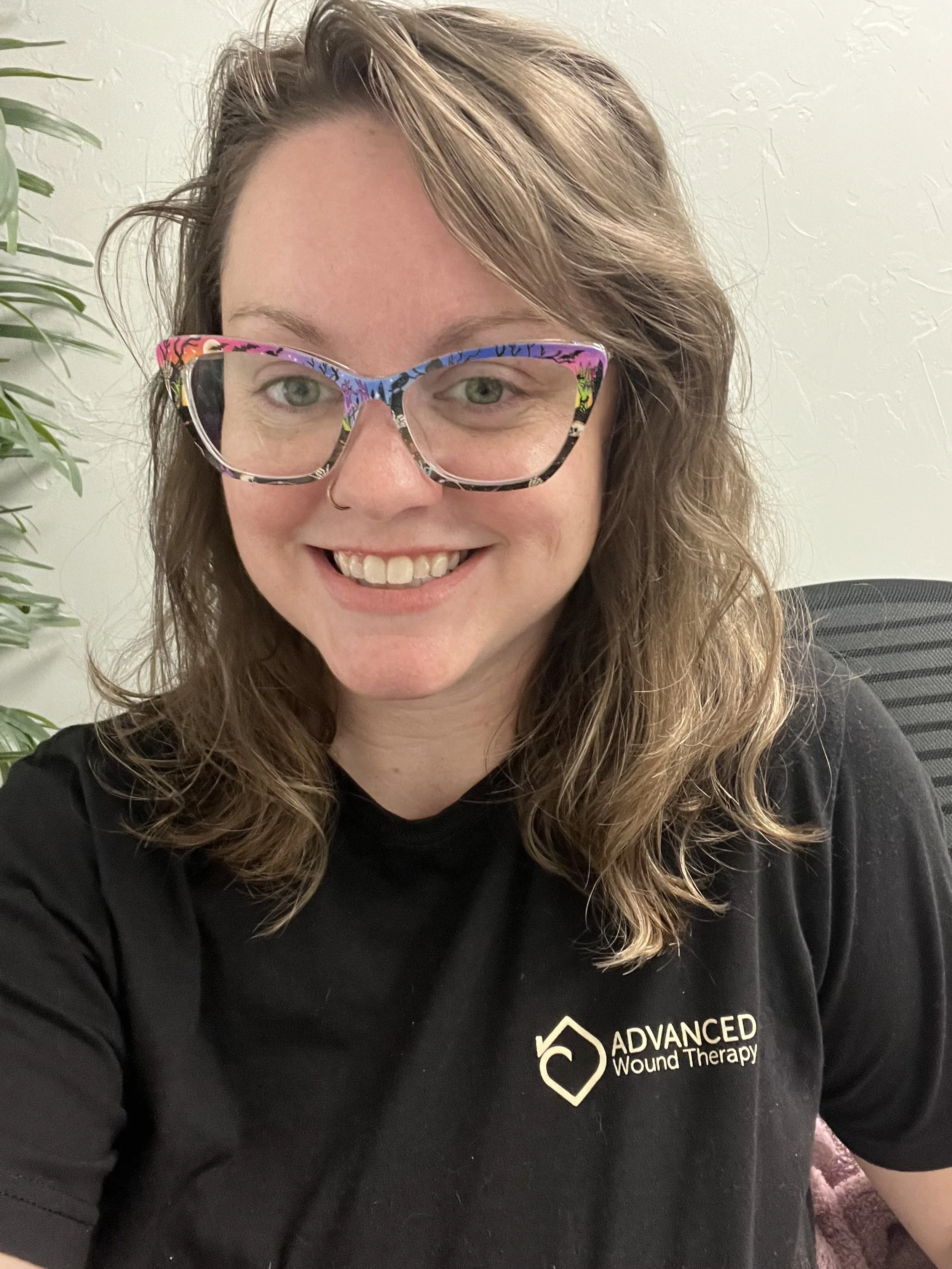 A woman with wavy brown hair, wearing colorful, patterned glasses and a black T-shirt with a logo that says "Advanced Wound Therapy." She is smiling and sitting in an office with a plant visible in the background.