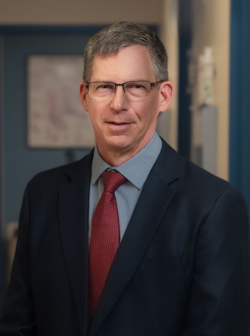 A middle-aged man wearing glasses, a dark suit, a light blue shirt, and a red tie, standing in an office setting.