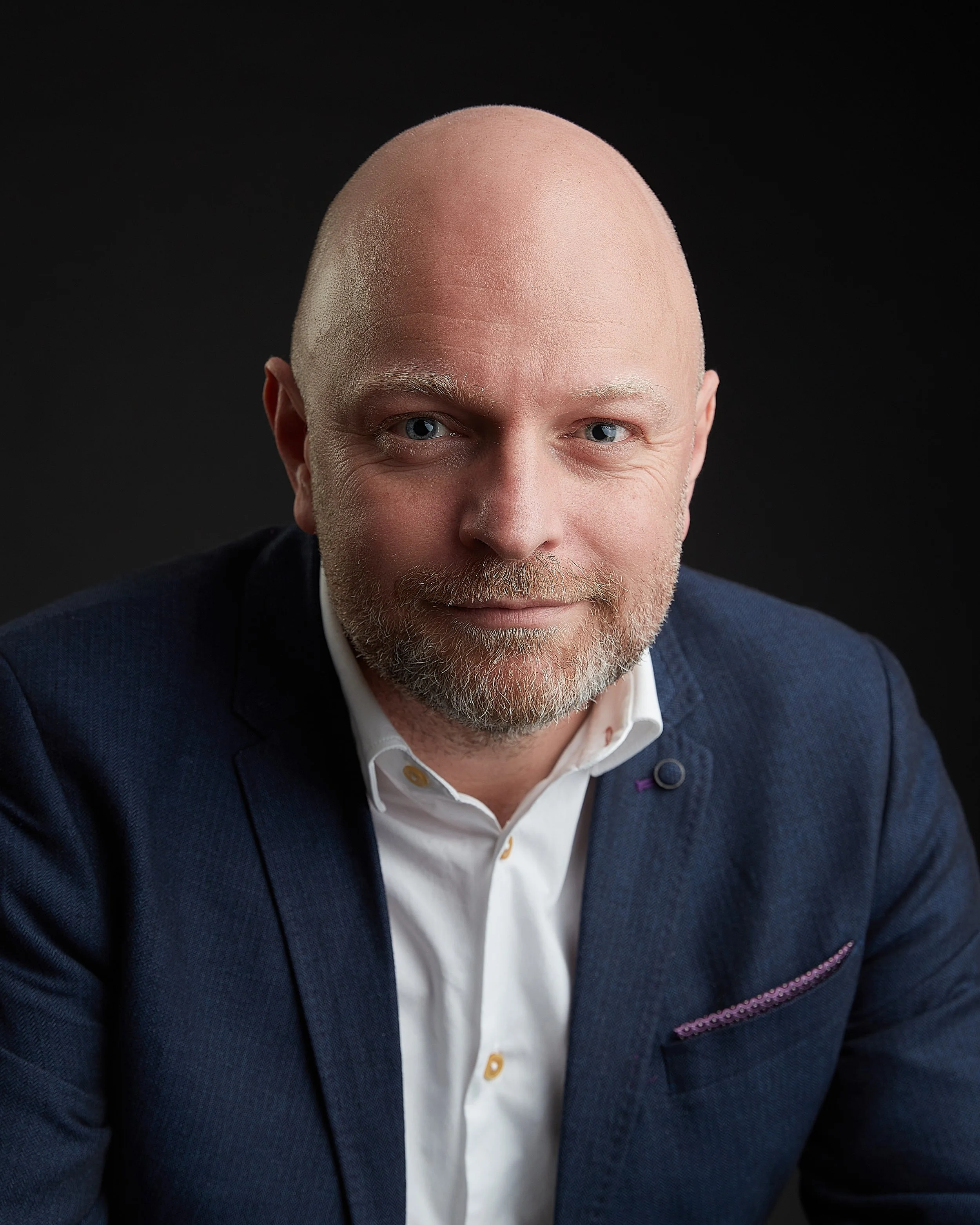 Headshot of a bald man with a gray beard wearing a navy blazer and white shirt against a black background.