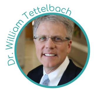 A portrait of a middle-aged man with glasses, short gray hair, wearing a dark suit, white shirt, and light blue tie, smiling against a blurred background.