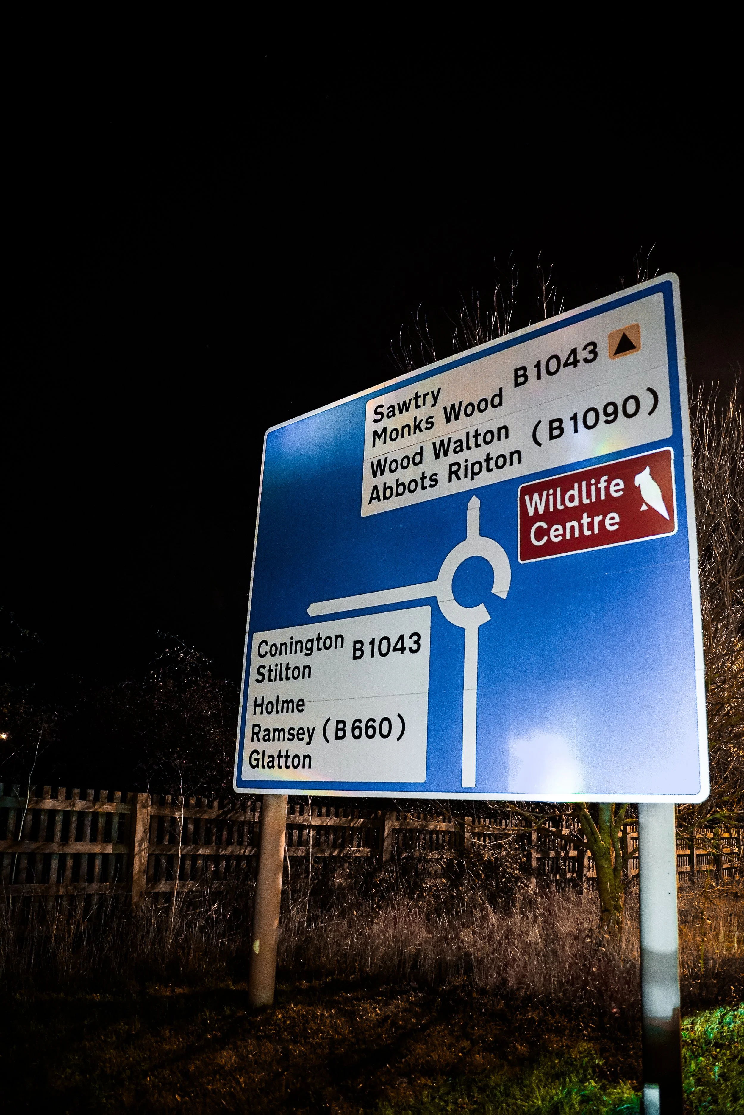 Nighttime road sign indicating directions to Sawtry, Monks Wood, Wood Walton, Abbots Ripton, Conington, Stilton, Holme, Ramsey, and Glatton, with a wildlife center nearby.