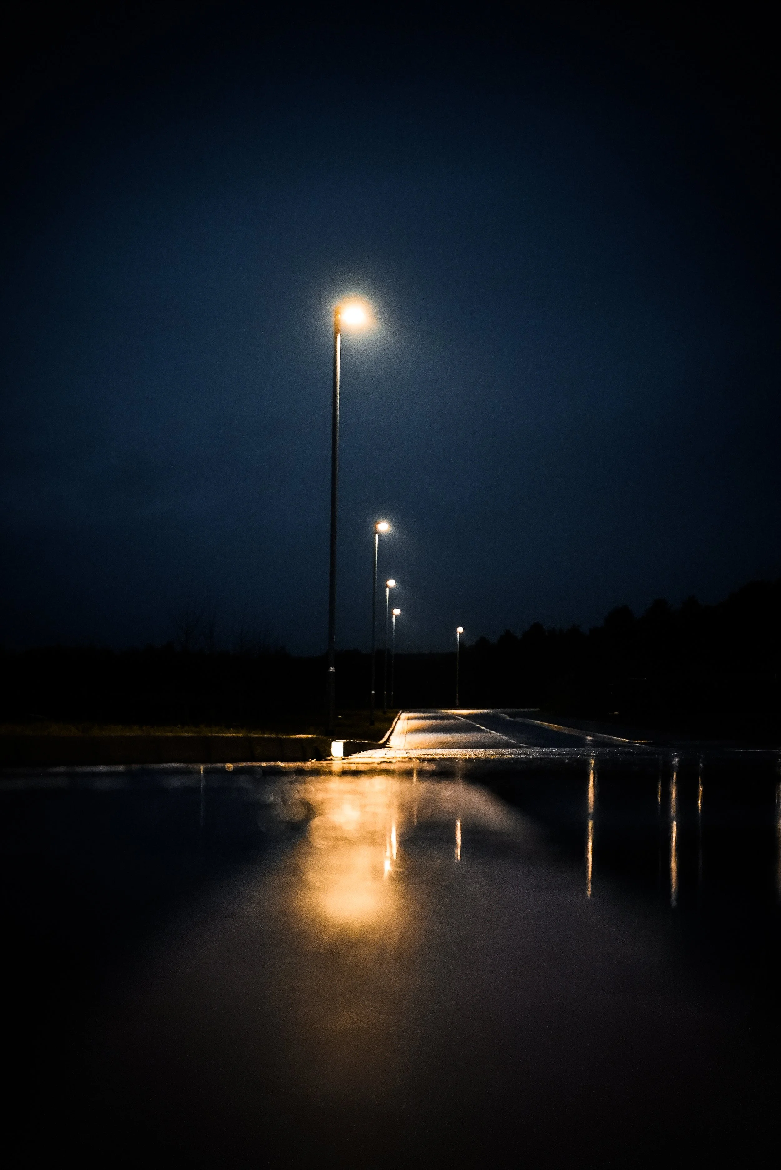 Nighttime view of a dimly lit street with several street lamps casting reflections on the wet pavement, stretching into the distance with a dark sky overhead.