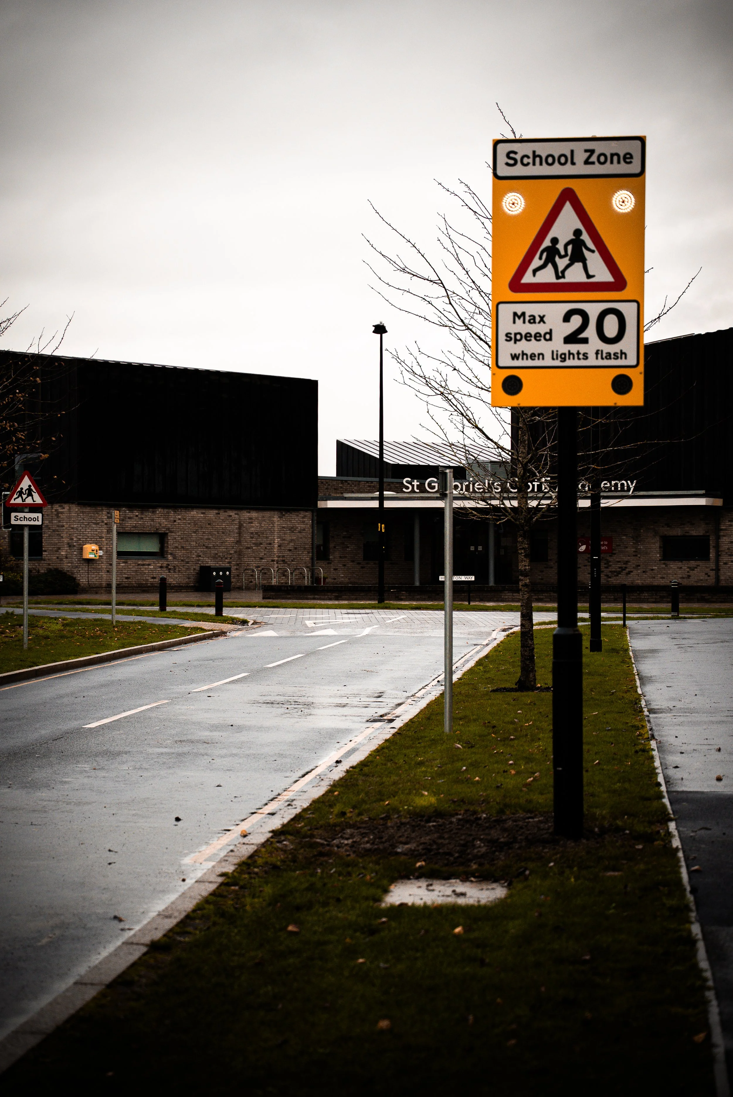 A school zone traffic sign indicating a maximum speed of 20 miles per hour when lights flash, with another smaller school zone sign in the background near a school entrance.