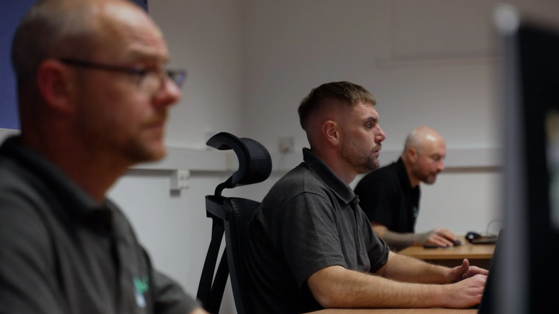 Three men sitting in a row at desks using computers, side profile view, in an office or classroom setting.