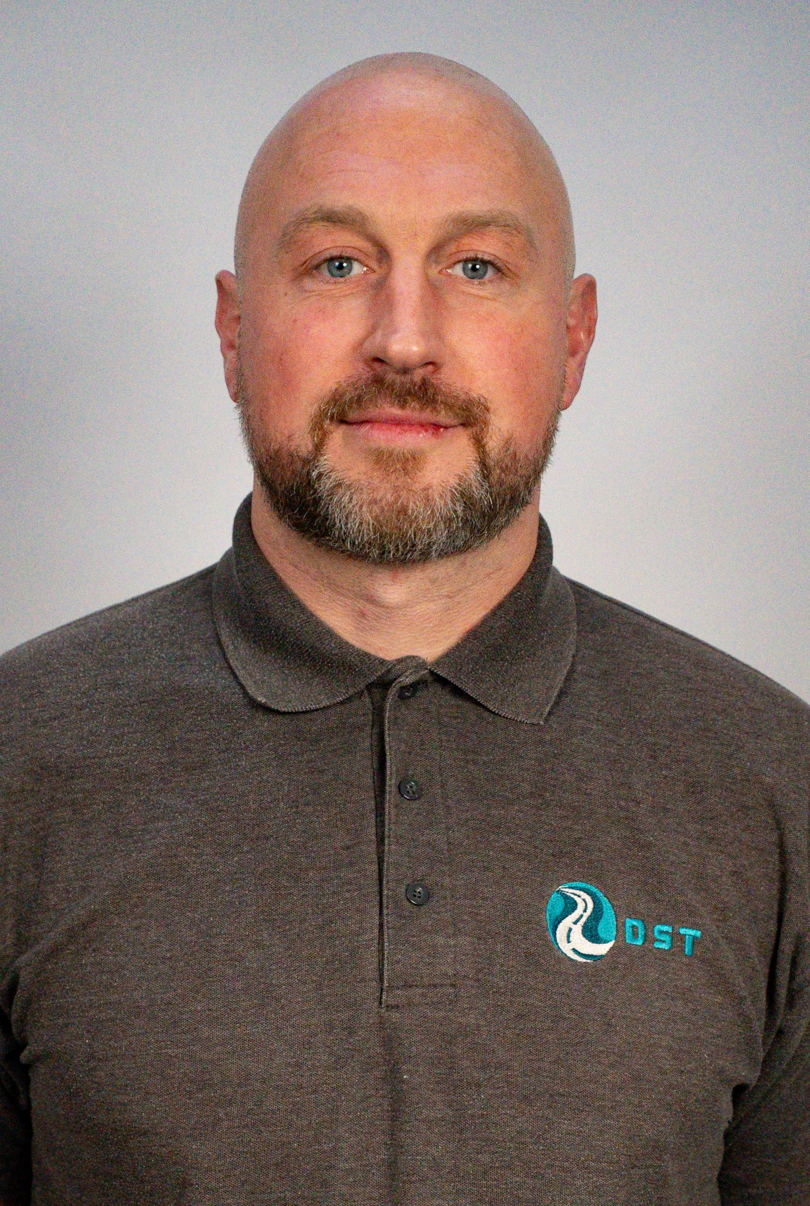 A middle-aged man with a beard and bald head, wearing a dark gray polo shirt with a logo on the left chest area that reads 'DST' alongside a wave-themed icon.