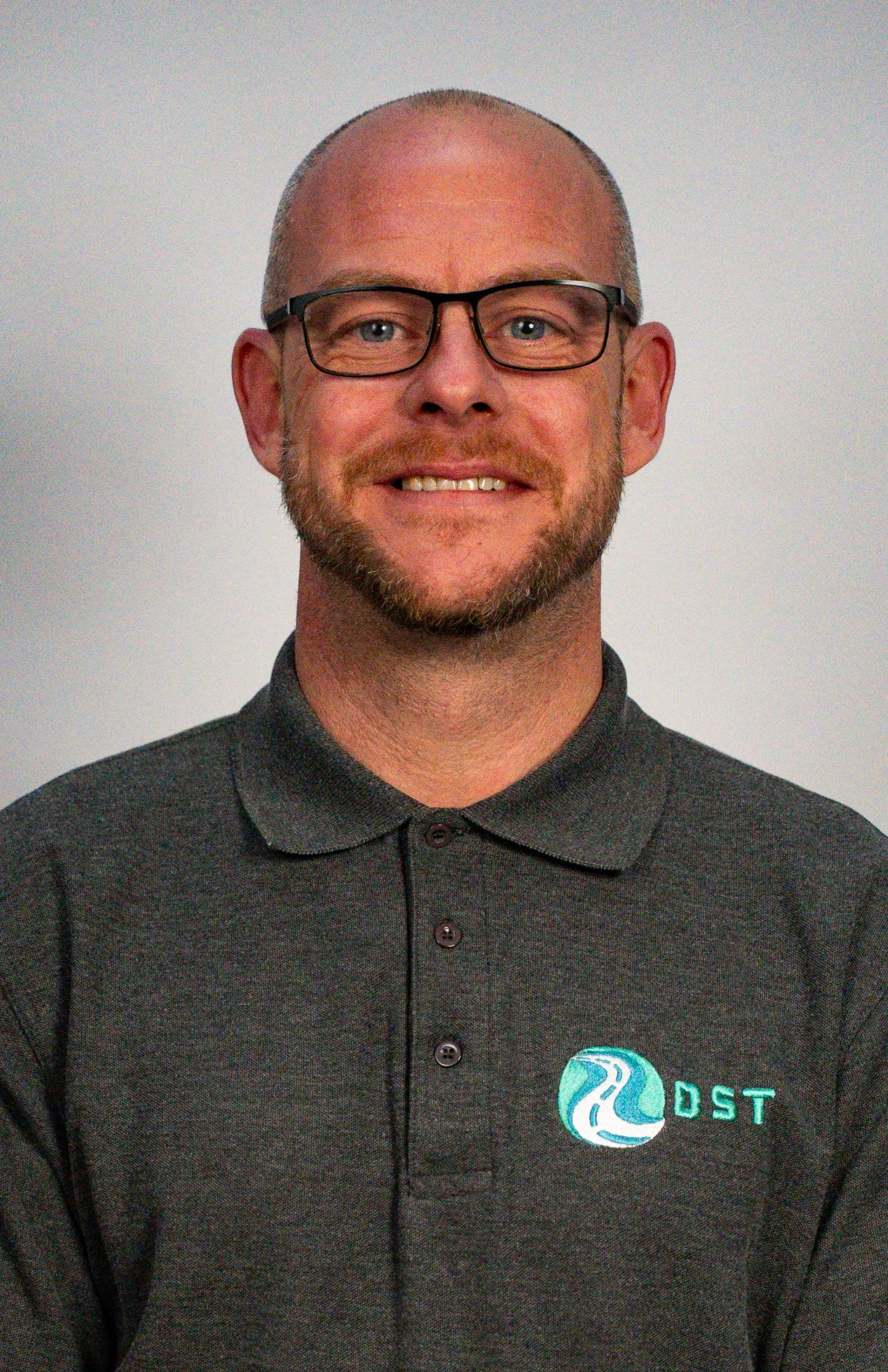 A man with glasses and a beard, wearing a black polo shirt with a logo that has a blue and white winding river and the letters "DST" on it, smiling in front of a plain background.