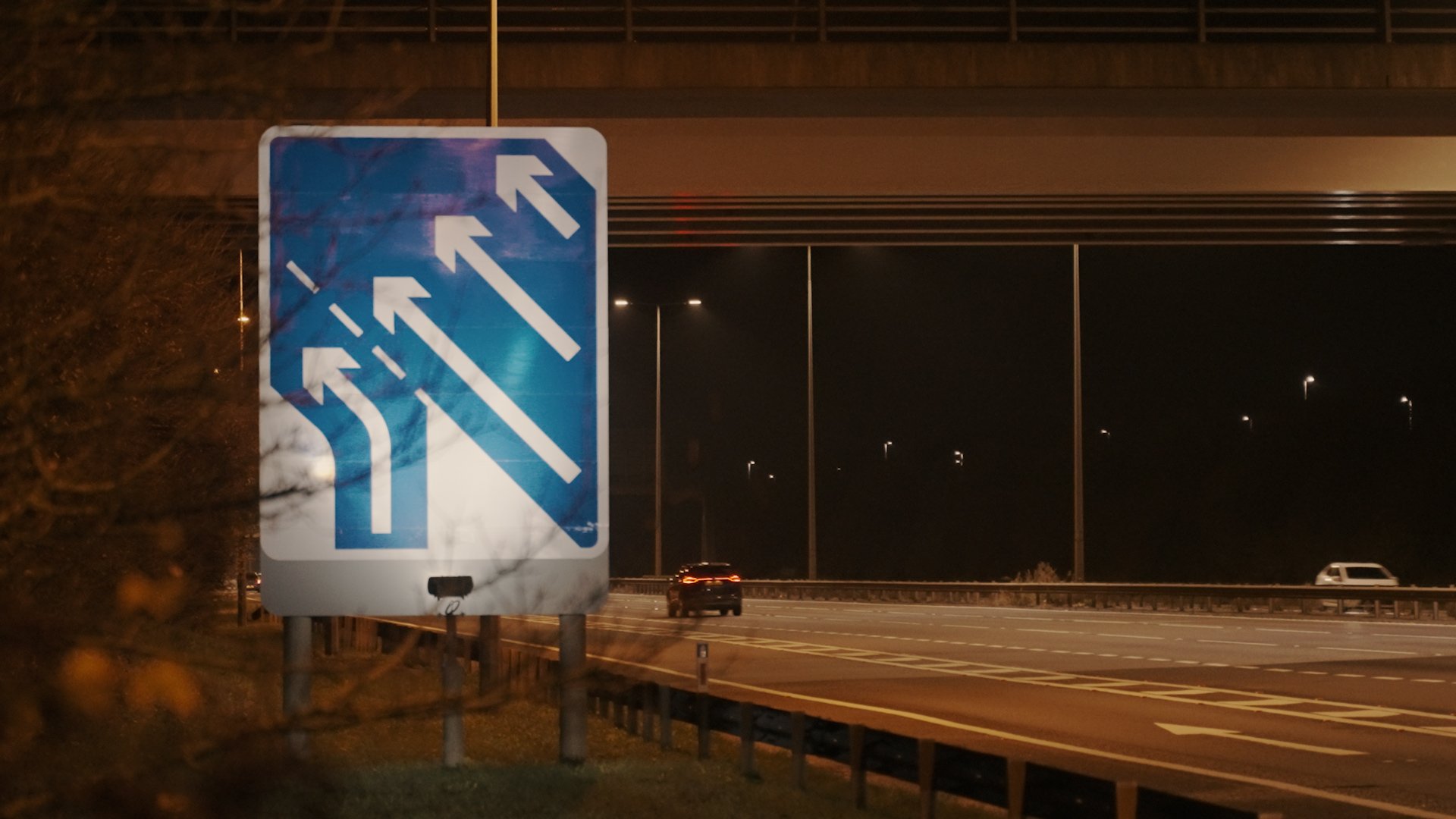 Nighttime highway scene with a sign showing three lanes moving to the left. Vehicles are visible on the road. Overpass is in the background.