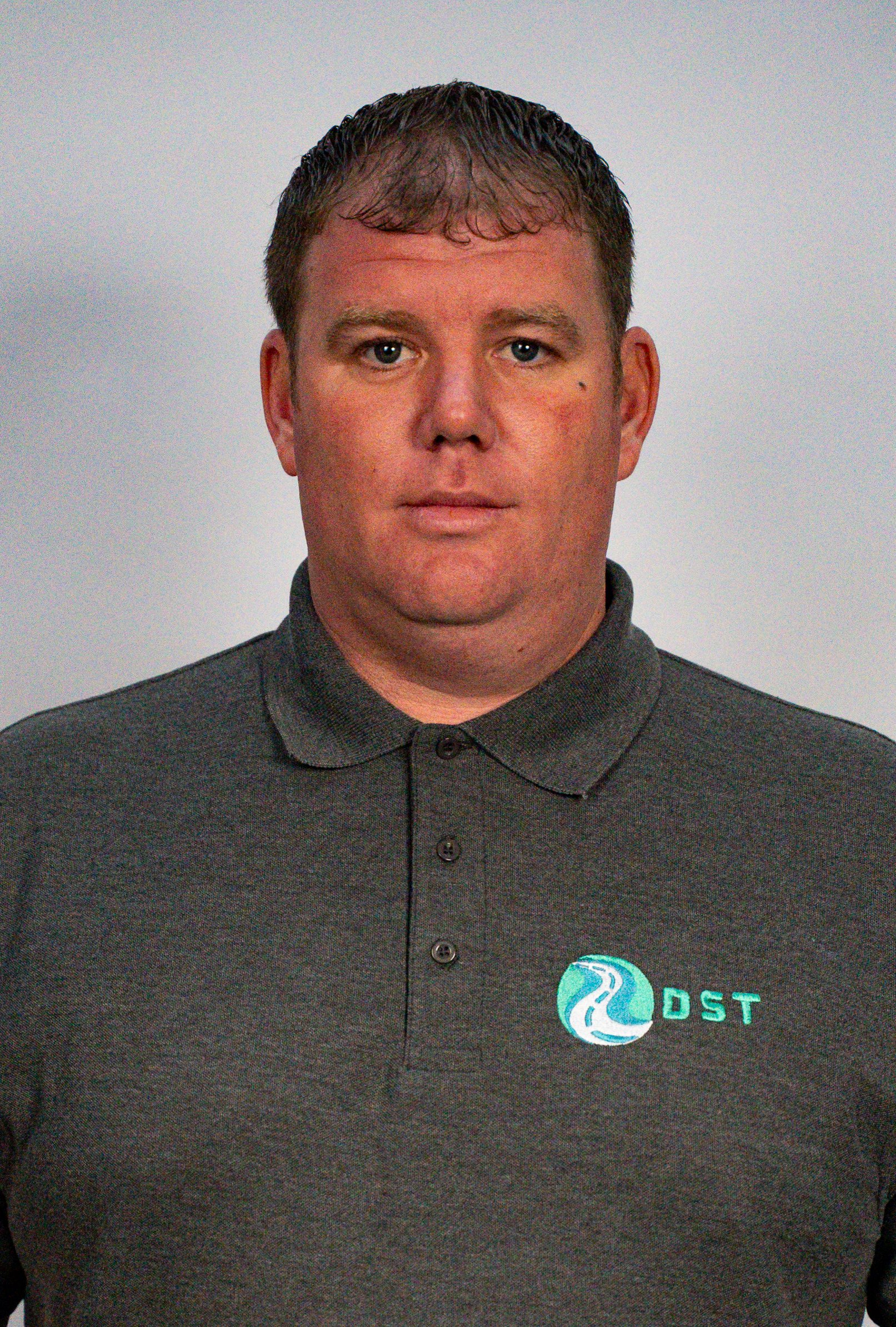 A man with short brown hair, wearing a dark gray polo shirt with a logo on the chest, standing against a plain light gray background.