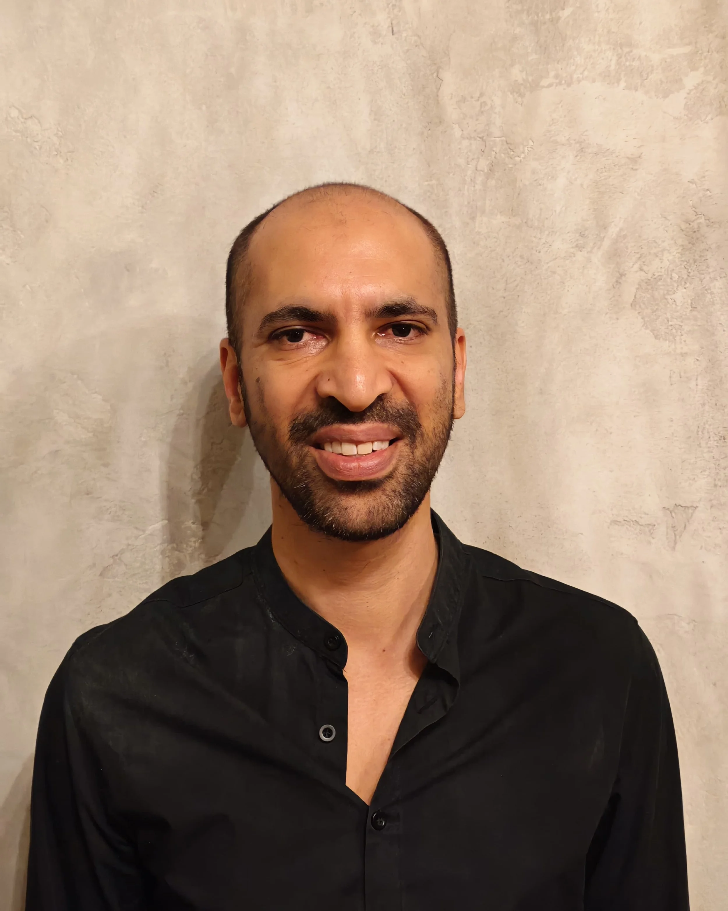A man with a short beard and bald head, wearing a black button-up shirt, smiling in front of a plain, textured beige wall.