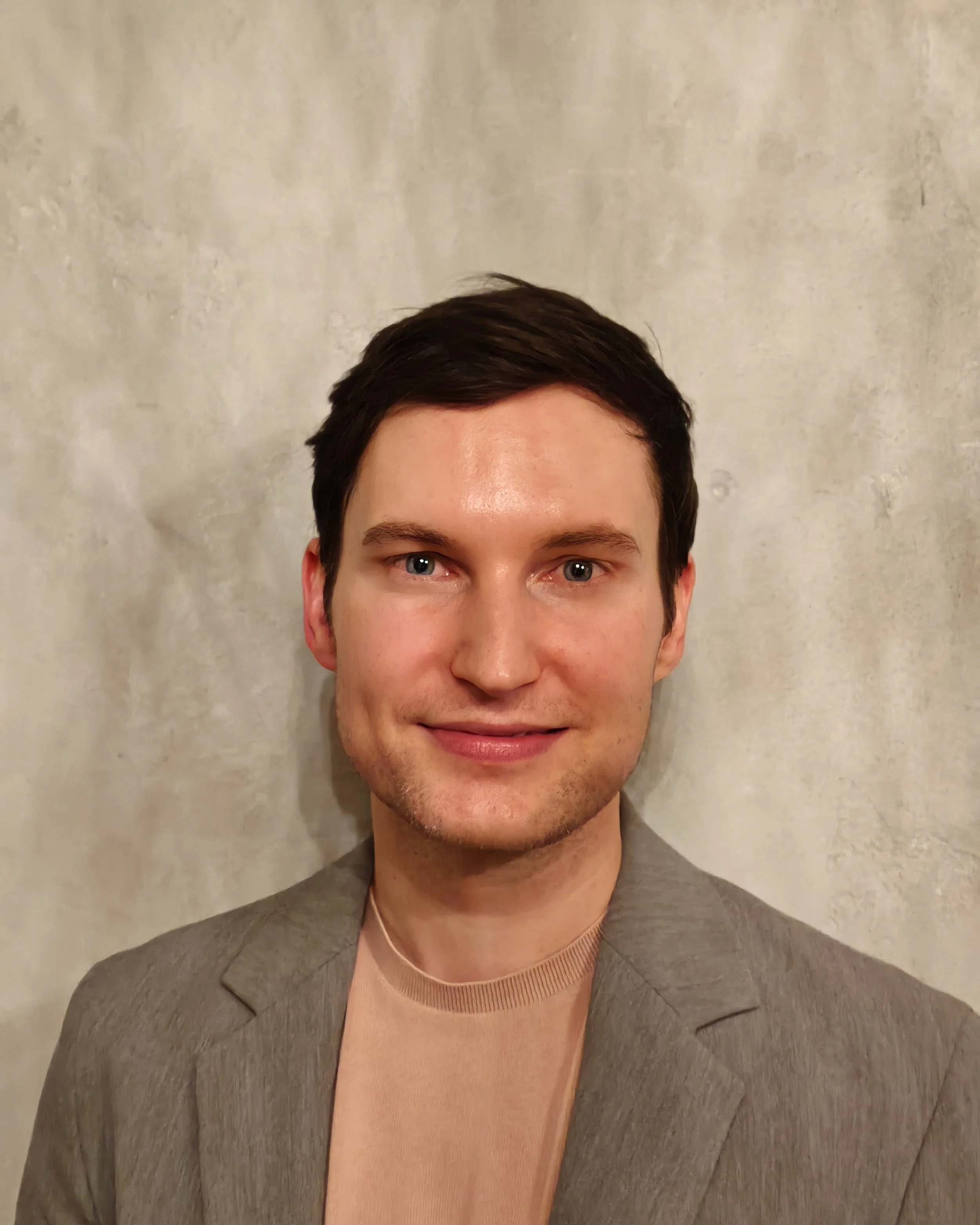 A young man with short dark hair, light skin, and blue eyes smiling at the camera, wearing a beige blazer and a pinkish-beige shirt against a textured light beige wall.