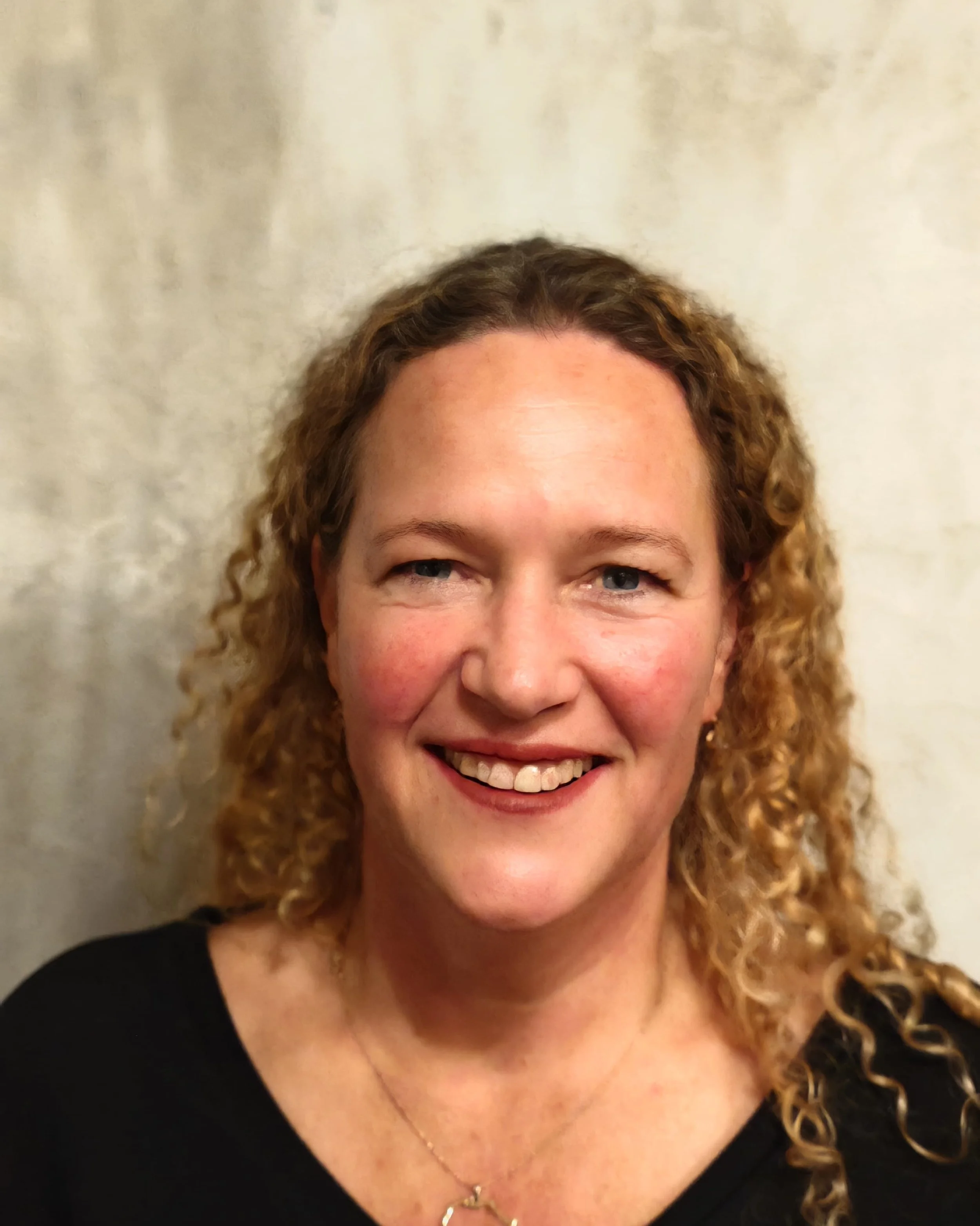 Close-up portrait of a smiling woman with curly blonde hair, wearing a black top and a necklace, posed against a light-colored textured background.