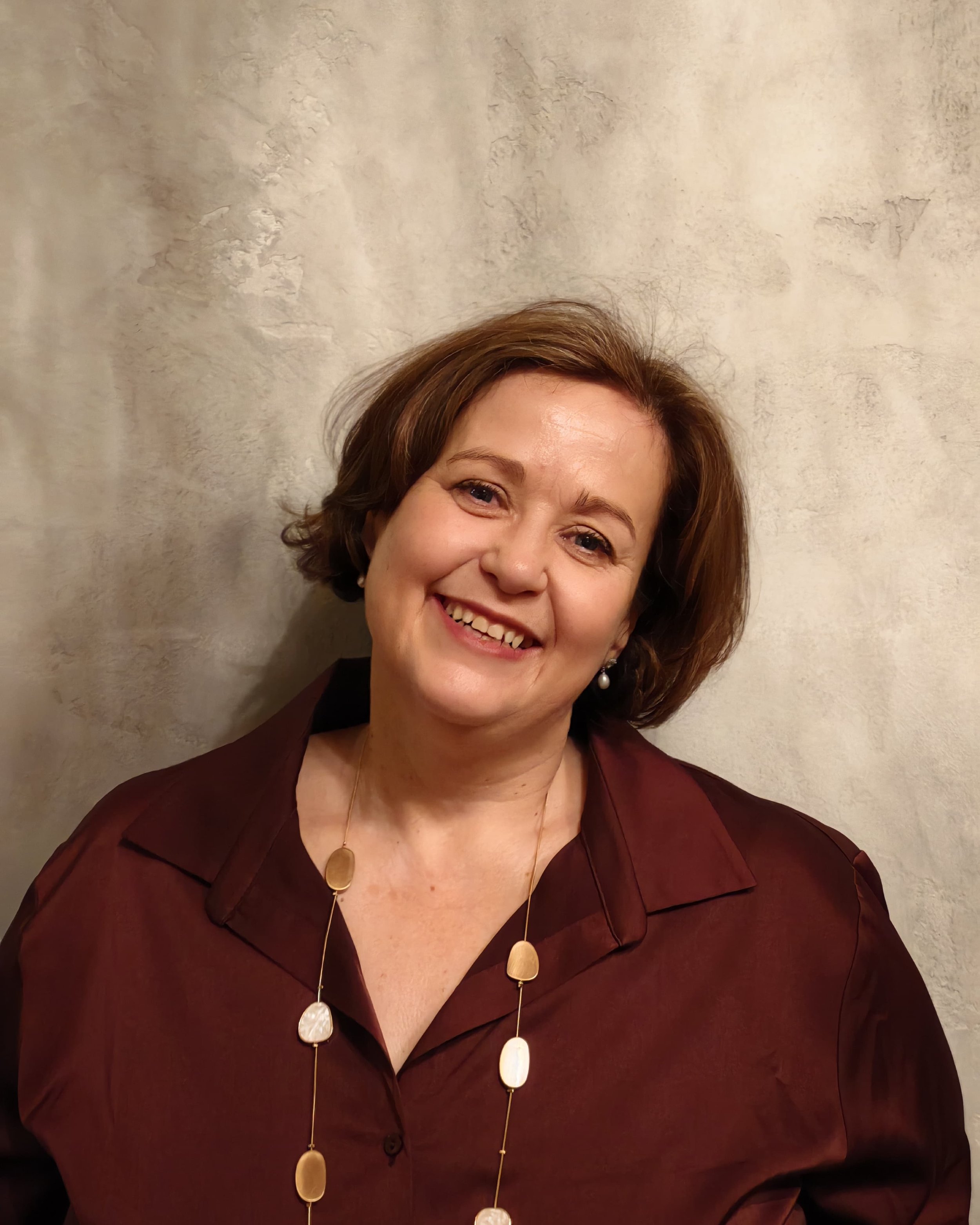 A smiling woman with short brown hair, wearing a maroon blouse and a necklace with oval pendants, standing against a textured beige wall.