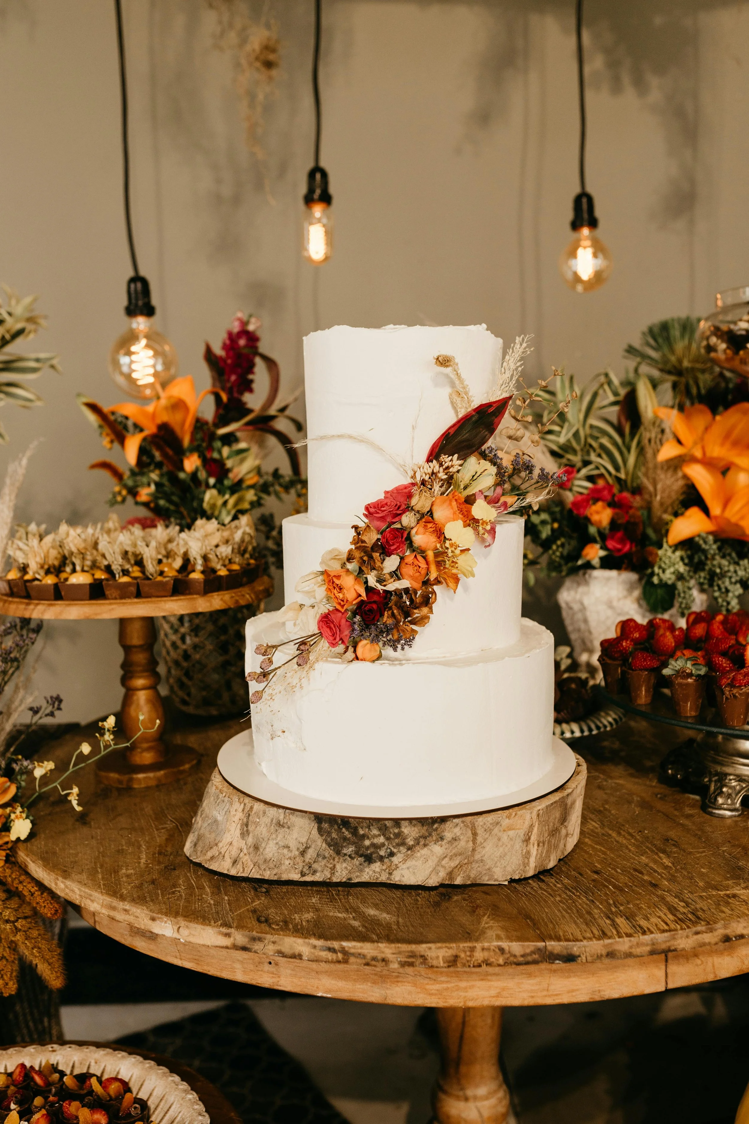 A three-tier white wedding cake decorated with a cascade of orange, red, and purple flowers, placed on a rustic wood slice on a wooden table.