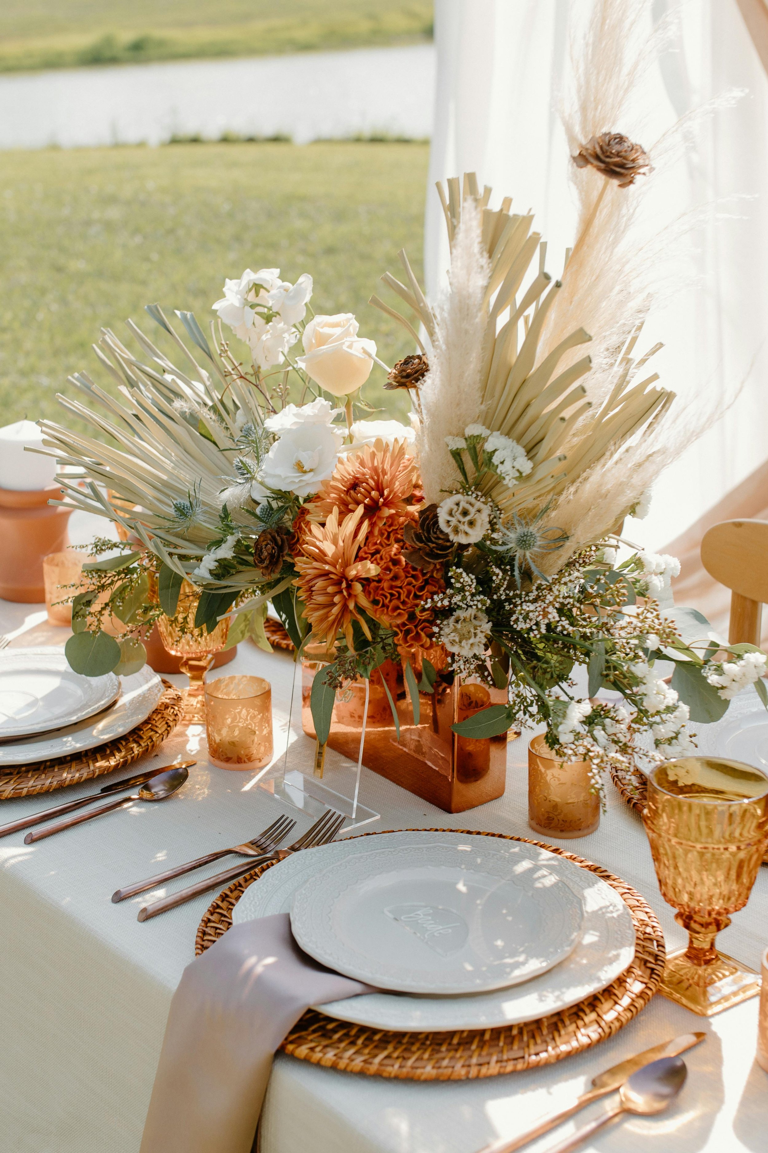 Elegant outdoor table setting with a floral centerpiece, white plates with a 'love' message, amber glassware, and cutlery on a white tablecloth overlooking a scenic lakeside.