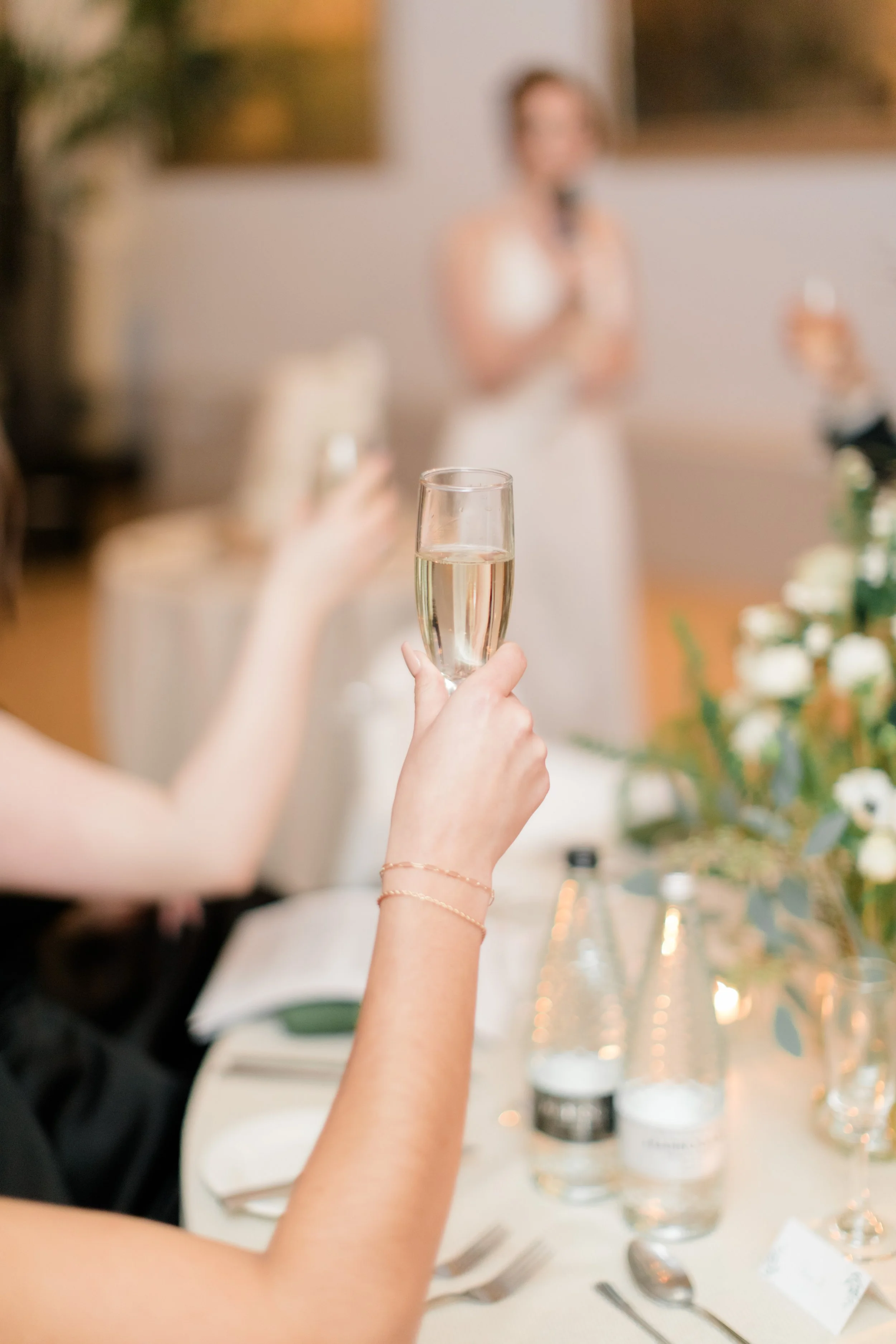 A person holding a glass of champagne at a wedding with women giving a speech in the background.