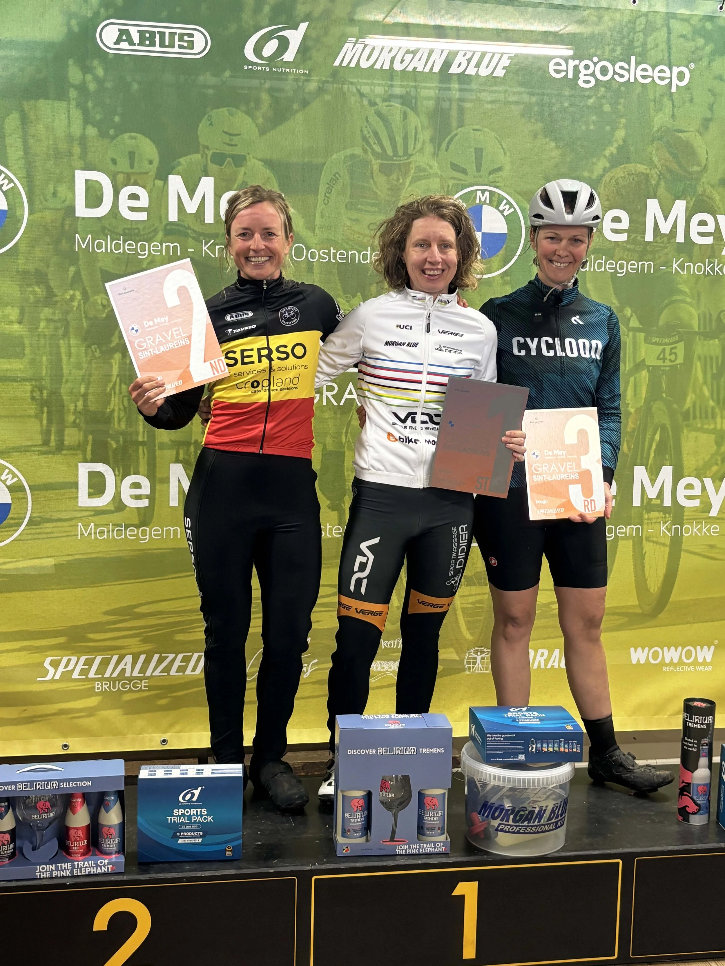 Drie vrouwelijke fietsers op een podium met prijzen en sponsorbanner.