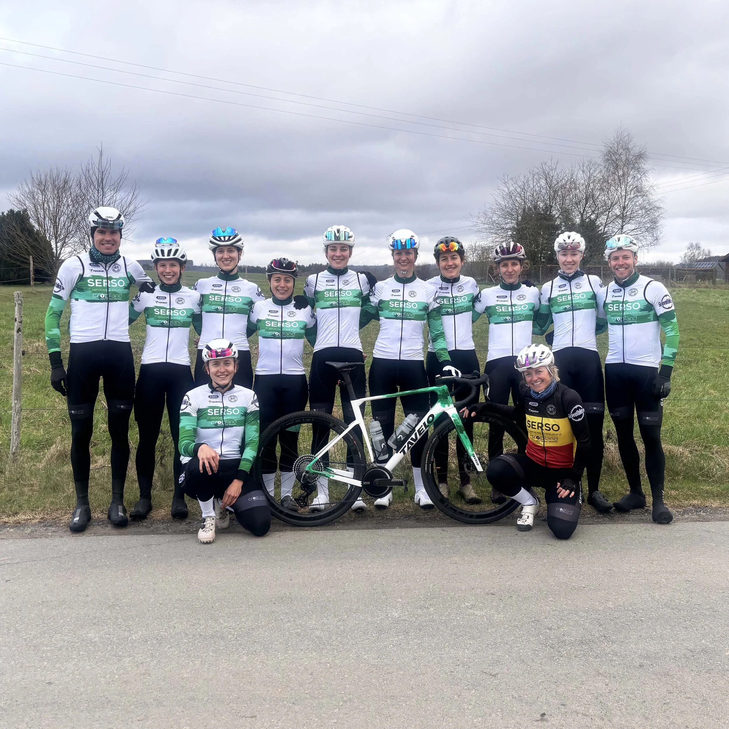 Een groep van twaalf wielrenners in een veld, gekleed in witte en groene teamkleding, het SerSo Gravelbike Team.