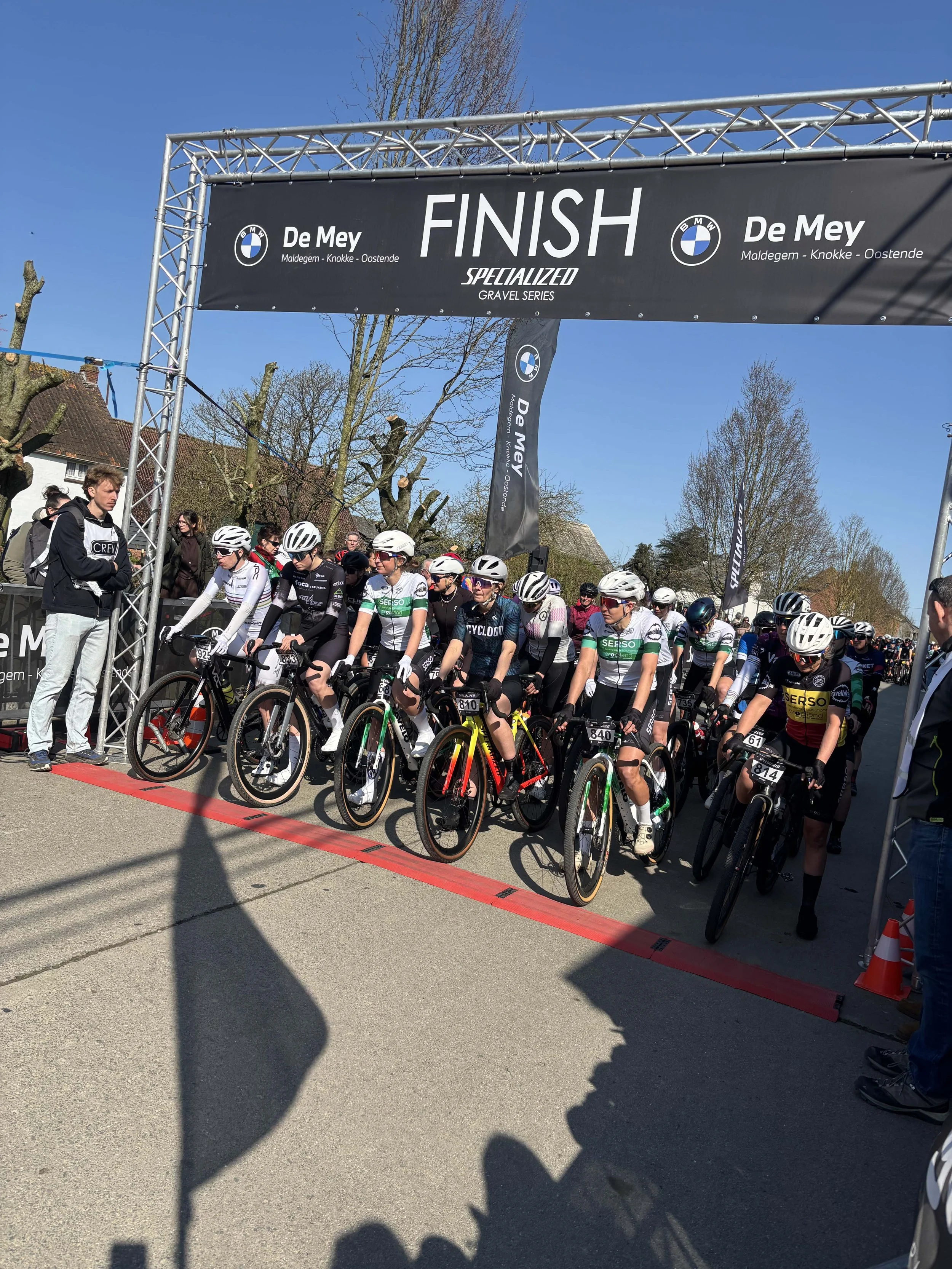 Wielrenners wachten aan de startlijn onder een finishboog bij een wielerevenement.