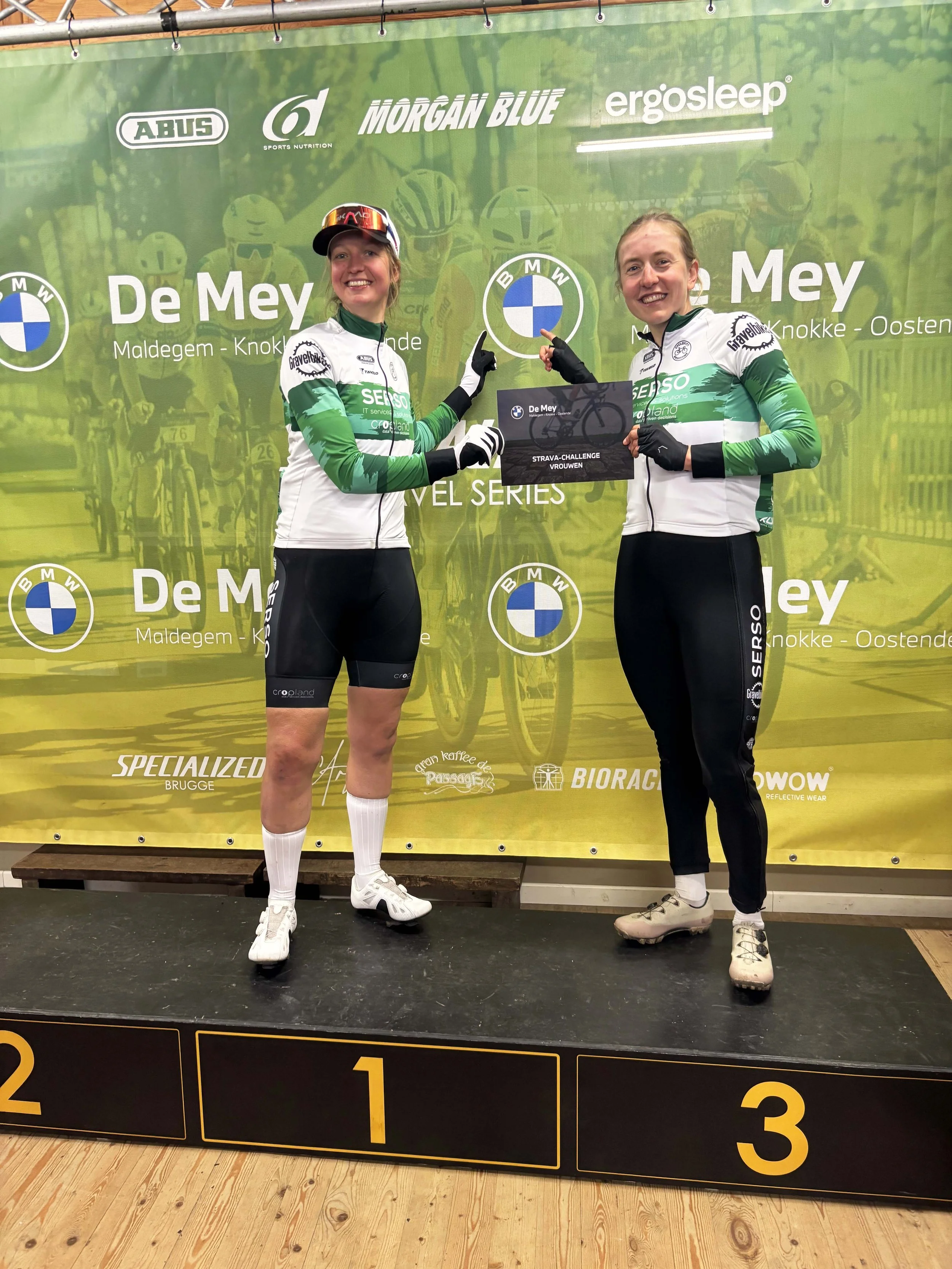 Twee wielrenners in wielerkleding staan op een podium, terwijl ze een prijs ontvangen. Beide vrouwen glimlachen en wijzen naar het bord dat aangeeft dat ze tweede en derde plaats hebben behaald. Op de achtergrond staat een groene banner met logo's en tekst.