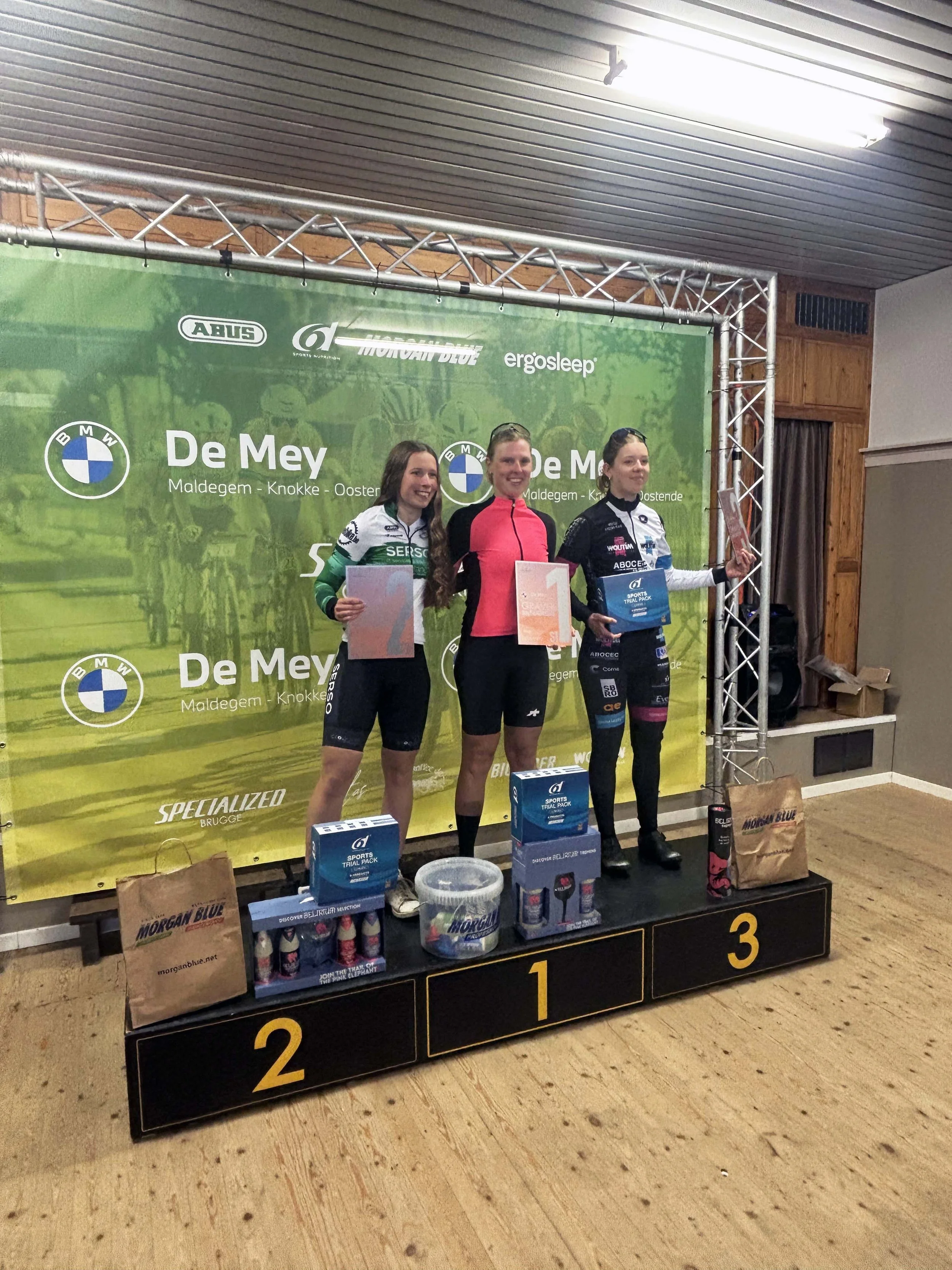 Drie vrouwen op het podium met prijzen op de eerste, tweede en derde plek, omringd door sporttassen en cadeaus, met een groene banner op de achtergrond.