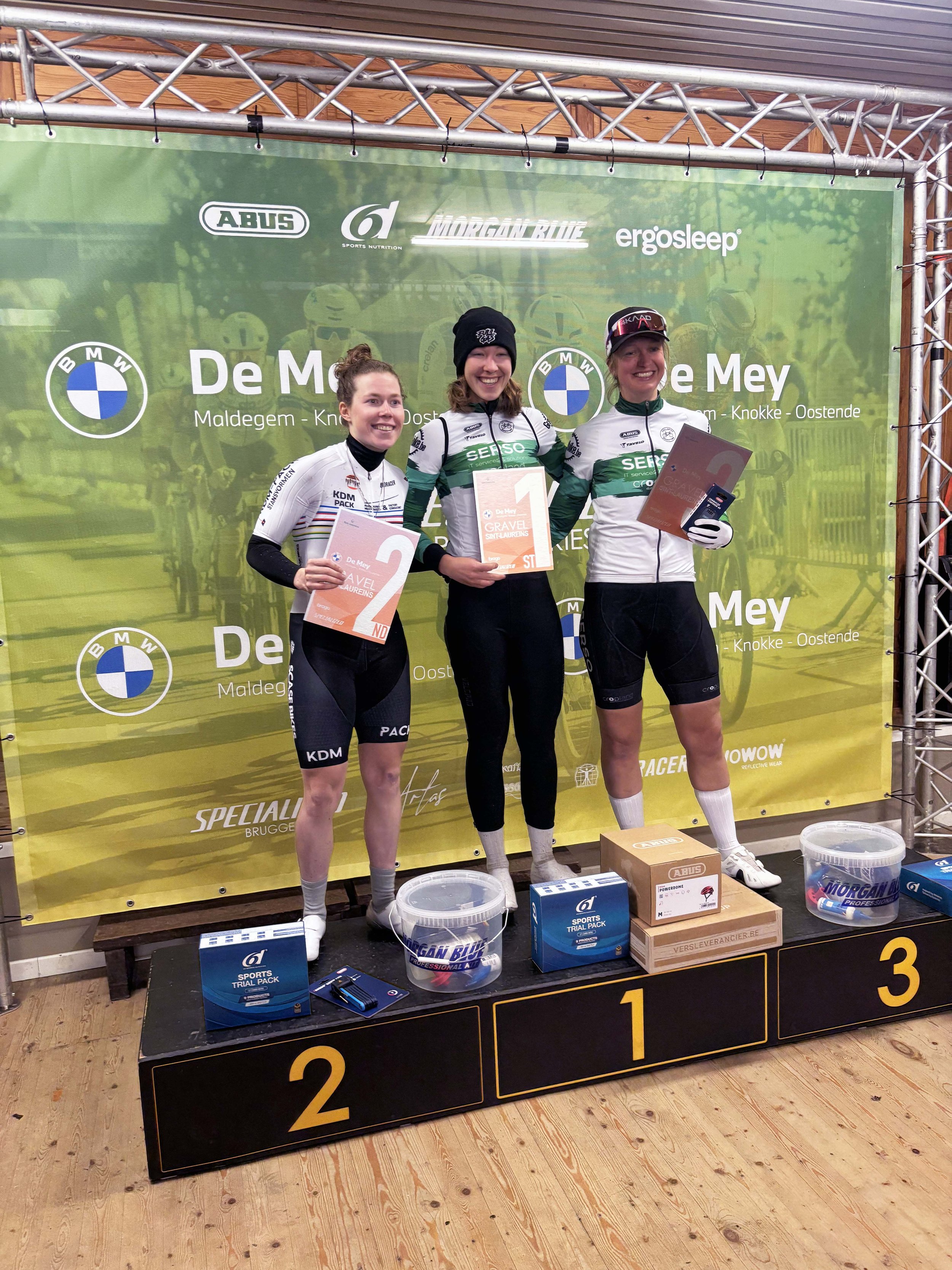 Drie vrouwelijke wielrenners op het podium, gekleed in wedstrijdkleding, poseren met prijzen en certificaten.