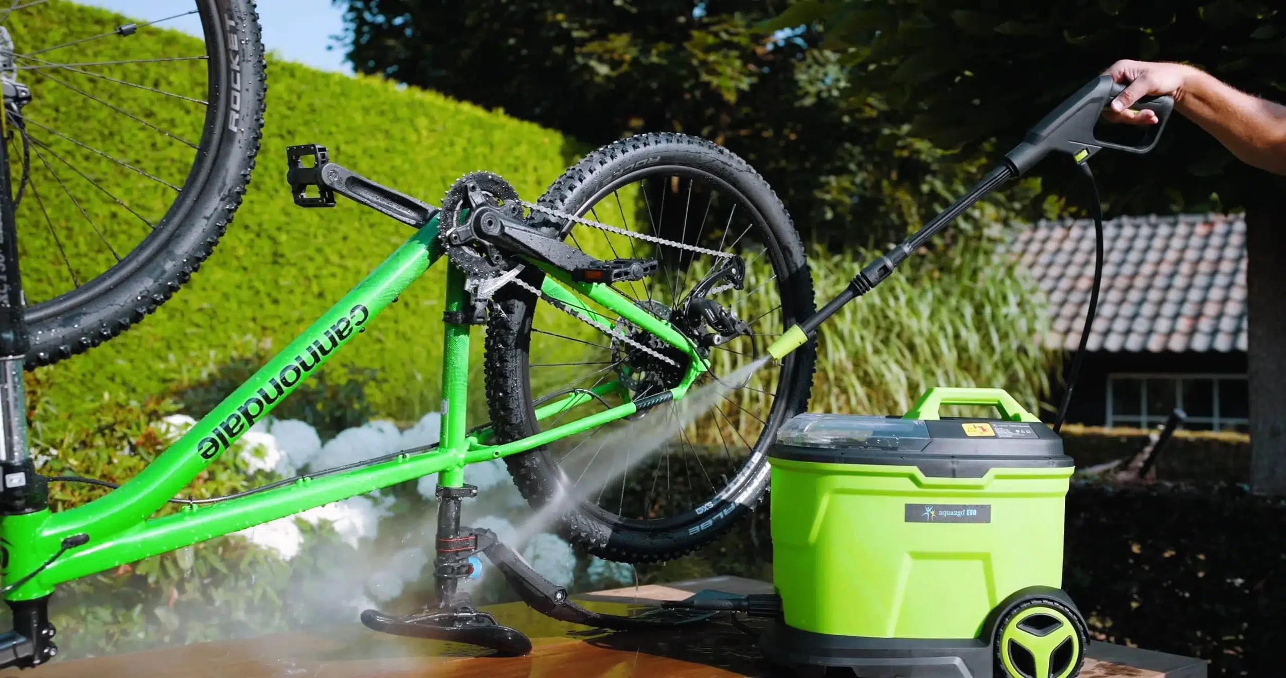 Een fietserreiniger die een groene mountainbike schoonmaakt met water onder een blauwe hemel. Er is groen gras en een boom in de achtergrond.